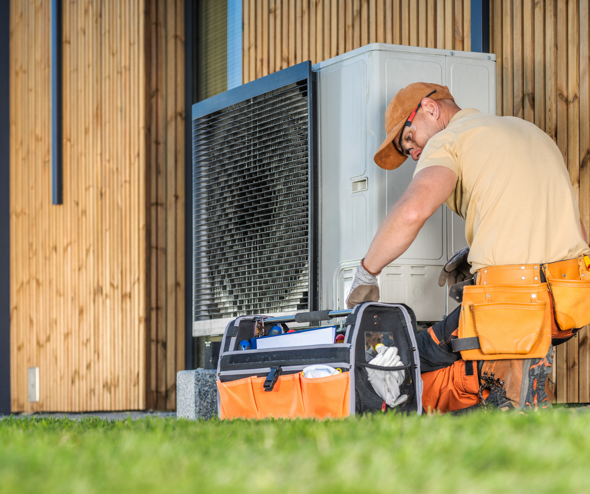 HVAC technician working on an outdoor unit. Orange tool belt, tools, brown cap, outside near a wooden wall.