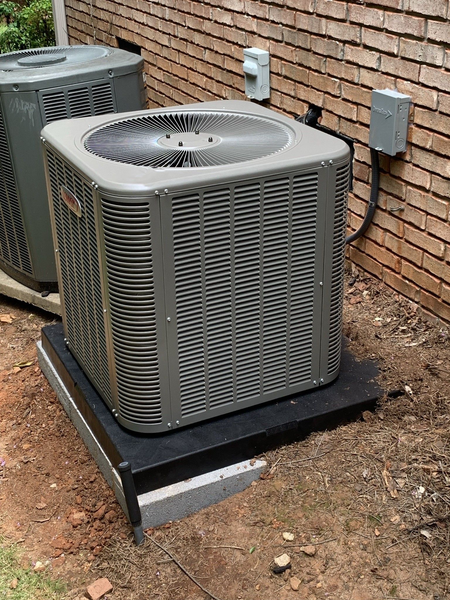 Air conditioning unit on a dark gray platform next to a brick wall. Electrical boxes are mounted on the wall.