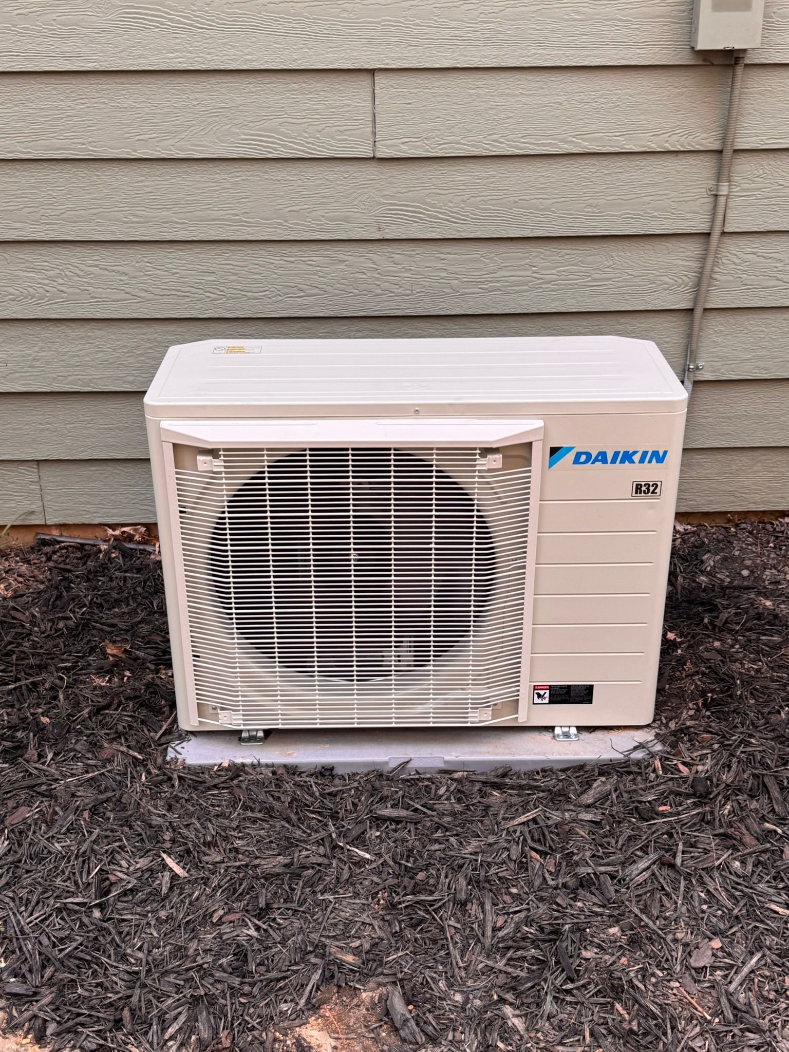 Daikin air conditioner unit against a siding wall, set on a concrete pad, surrounded by black mulch.