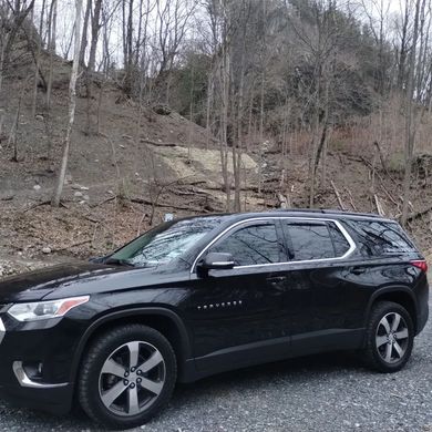 A black suv is parked in a gravel lot in front of a forest.