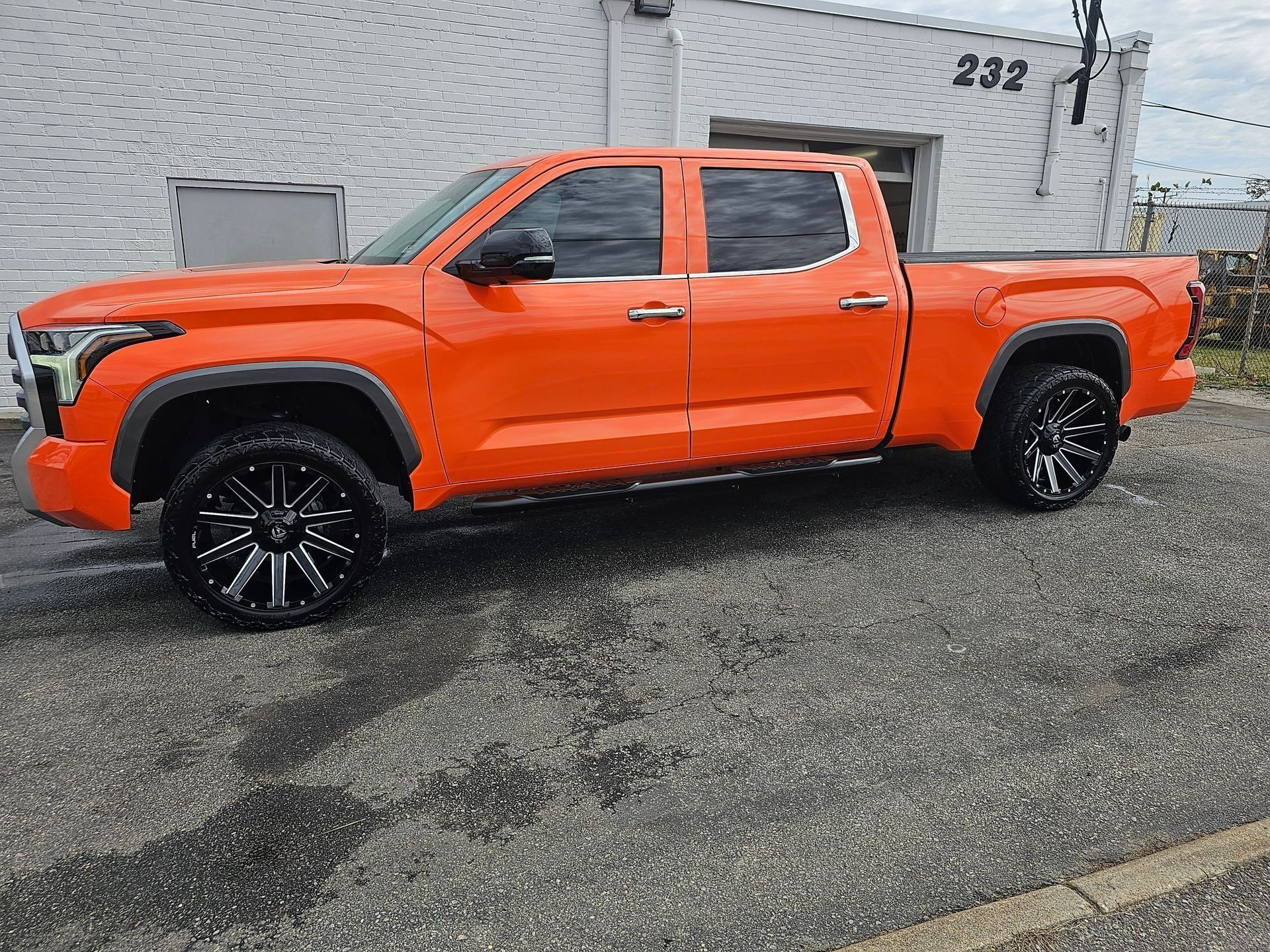 A bright orange crew cab pickup truck parked on a gravel lot in front of a white building marked with the number 232.