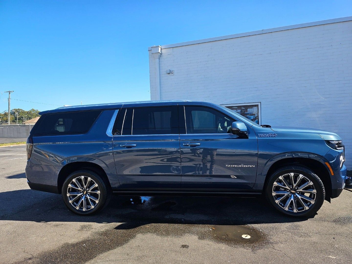 A blue Chevrolet Suburban parked in a paved lot next to a white brick wall under a clear blue sky.