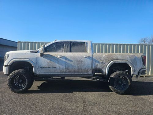 A muddy white pickup truck with oversized tires parked in front of a metal building under a clear blue sky.