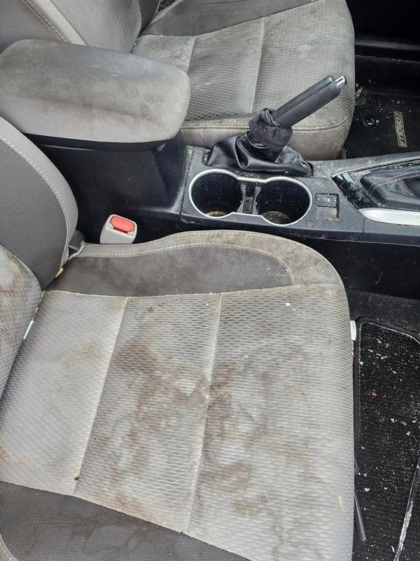 A car interior showing dirty gray cloth seats, a center console with two cup holders, and an upright parking brake handle.