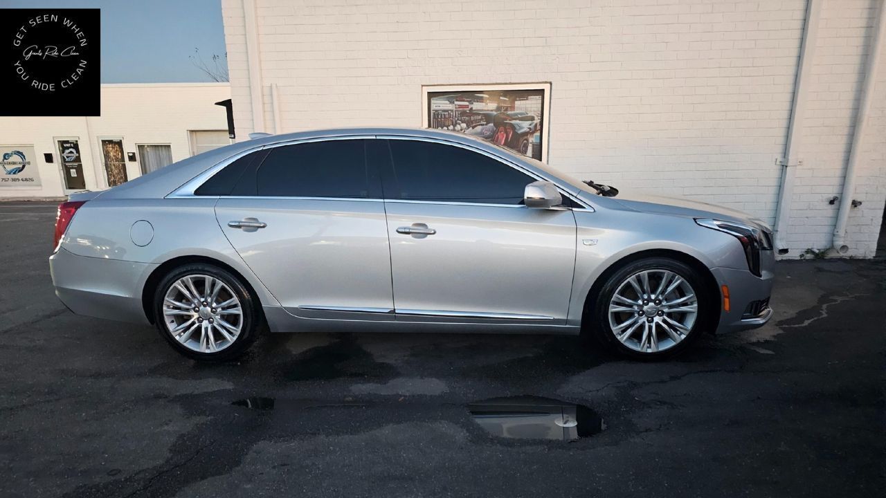 A silver Cadillac sedan parked on an asphalt lot beside a white brick building.