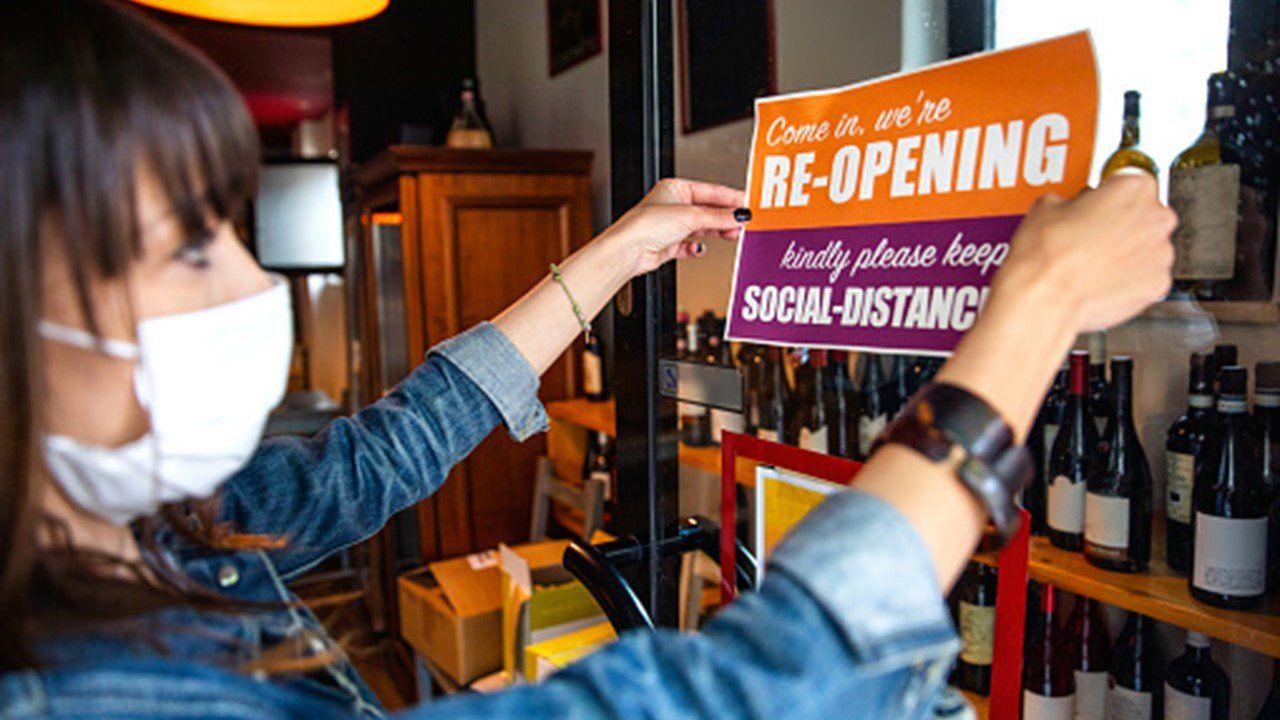 A woman wearing a mask is putting a reopening sign on a glass door.