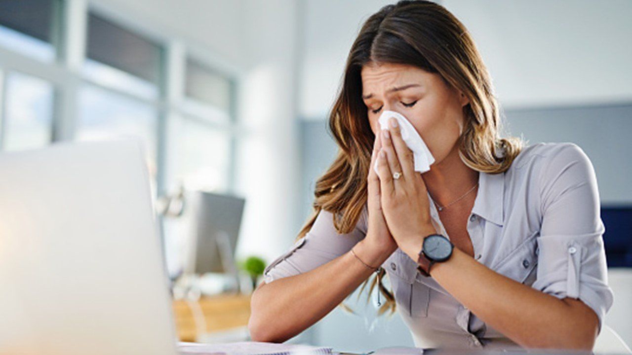 A woman is blowing her nose while sitting at a desk in front of a laptop computer.