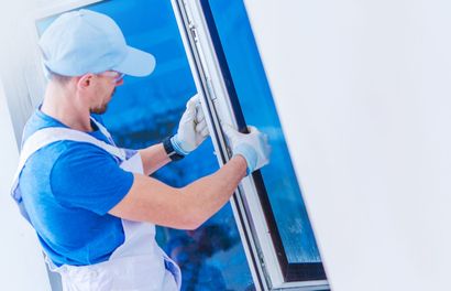 A technician wearing a blue shirt and white apron installs or repairs a white-framed window.