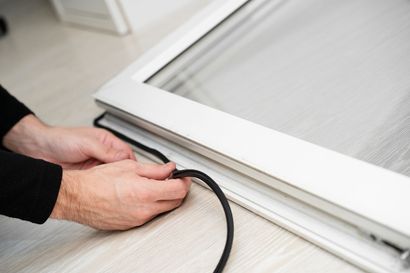 A pair of hands inserts a black rubber spline into the frame of a white window screen.