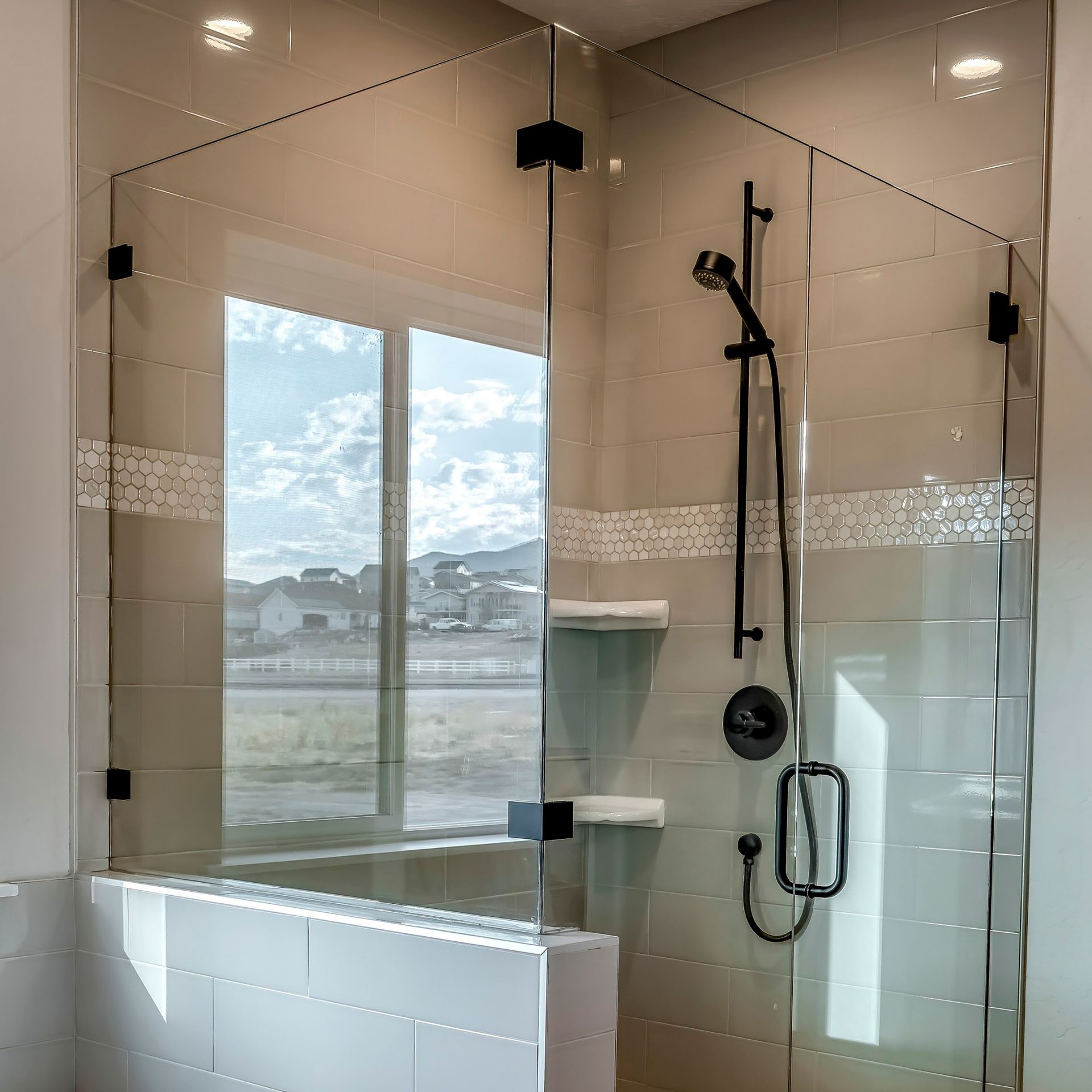 A modern glass shower enclosure with matte black hardware, installed on a tiled pony wall in a bright, sunlit bathroom.