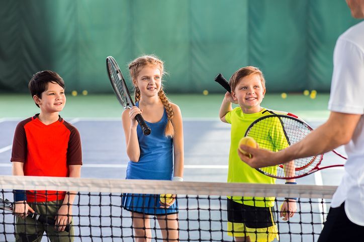A man is teaching three children how to play tennis on a tennis court.