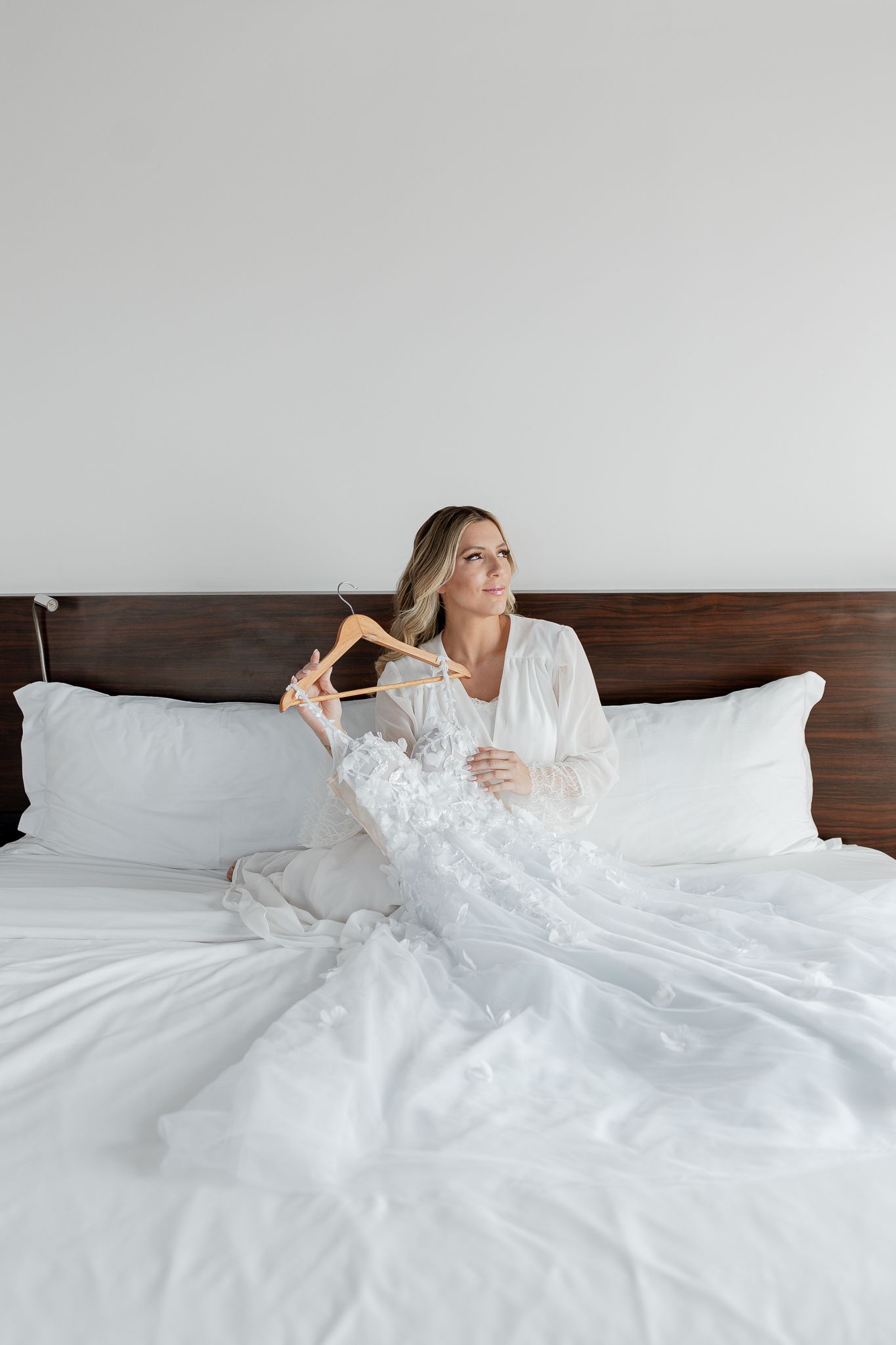 Mulher sentada na cama, segurando seu vestido de noiva branco. Ela tem cabelos loiros e olha para o lado.