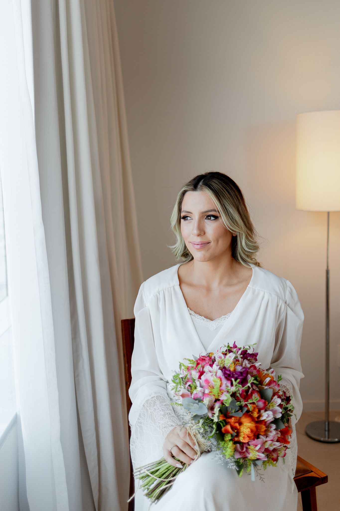 Mulher de vestido branco segurando um buquê colorido, sentada perto de uma janela. Ela tem cabelos loiros e parece pensativa.