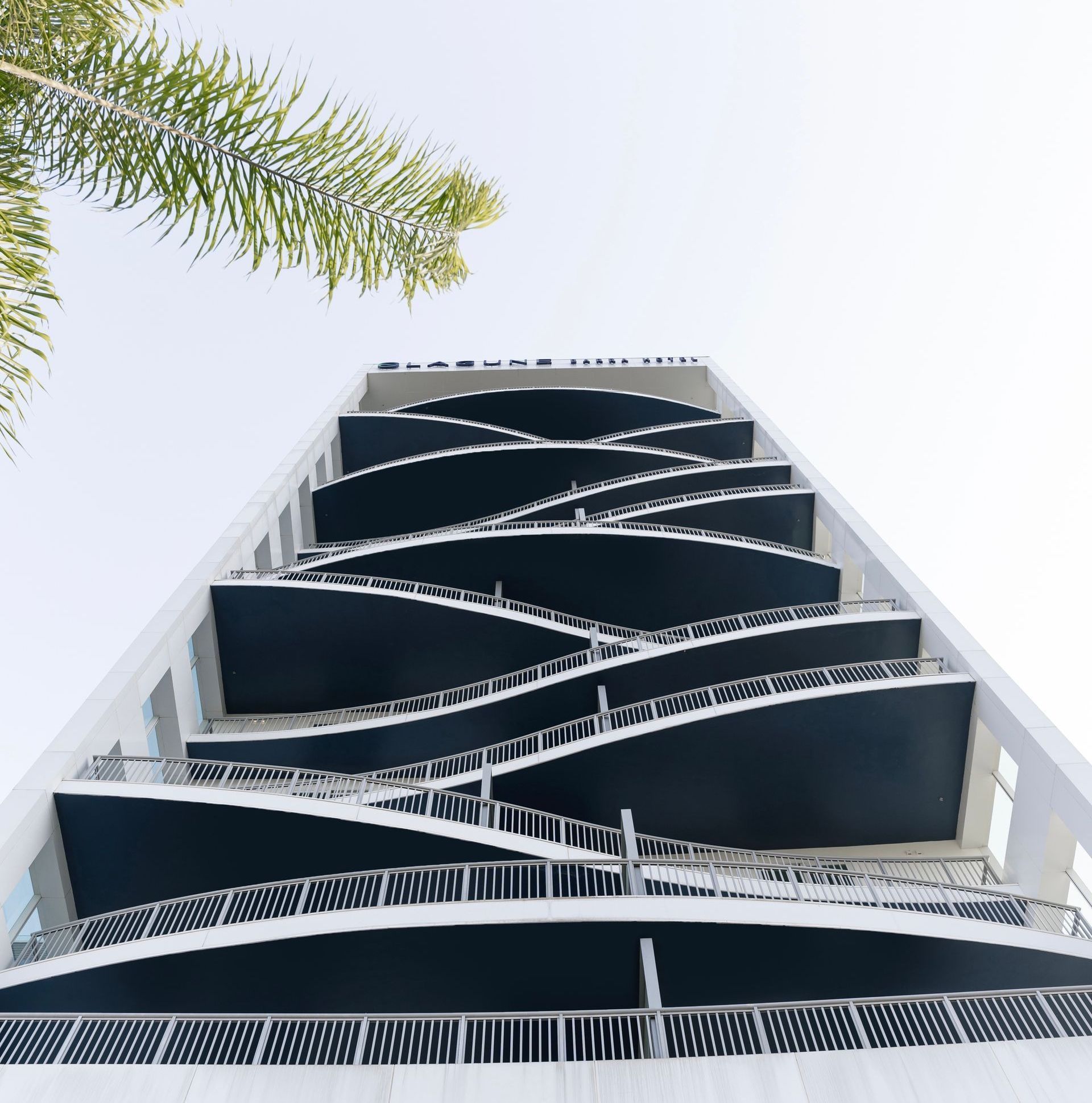 Vista de baixo para cima de um edifício branco moderno com varandas onduladas e escuras. Folhas de palmeira emolduram o canto superior esquerdo.