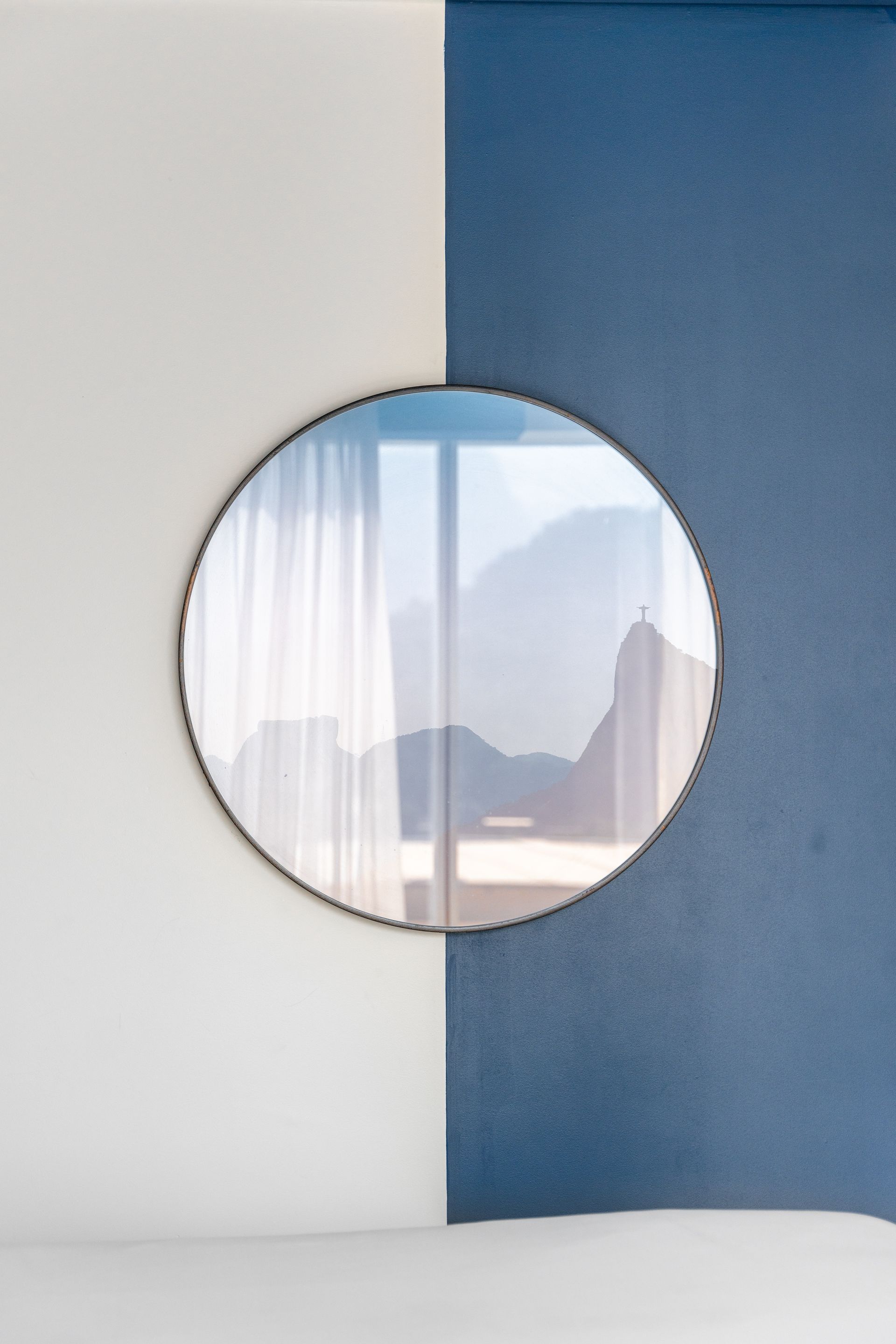 Espelho redondo refletindo a vista da montanha, montado em uma parede de dois tons (branco e azul).
