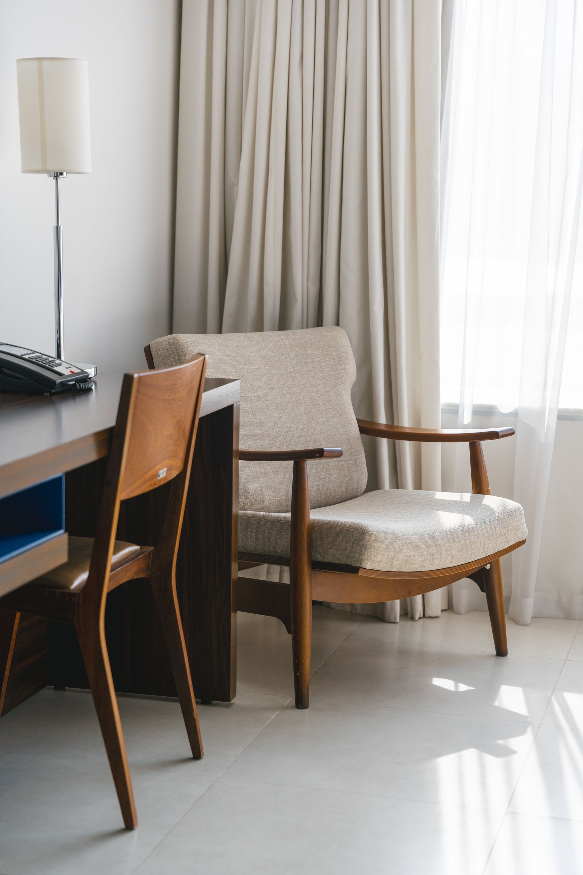 Um quarto de hotel em estilo moderno de meados do século, com escrivaninha, cadeira e poltrona perto de uma janela com cortinas.