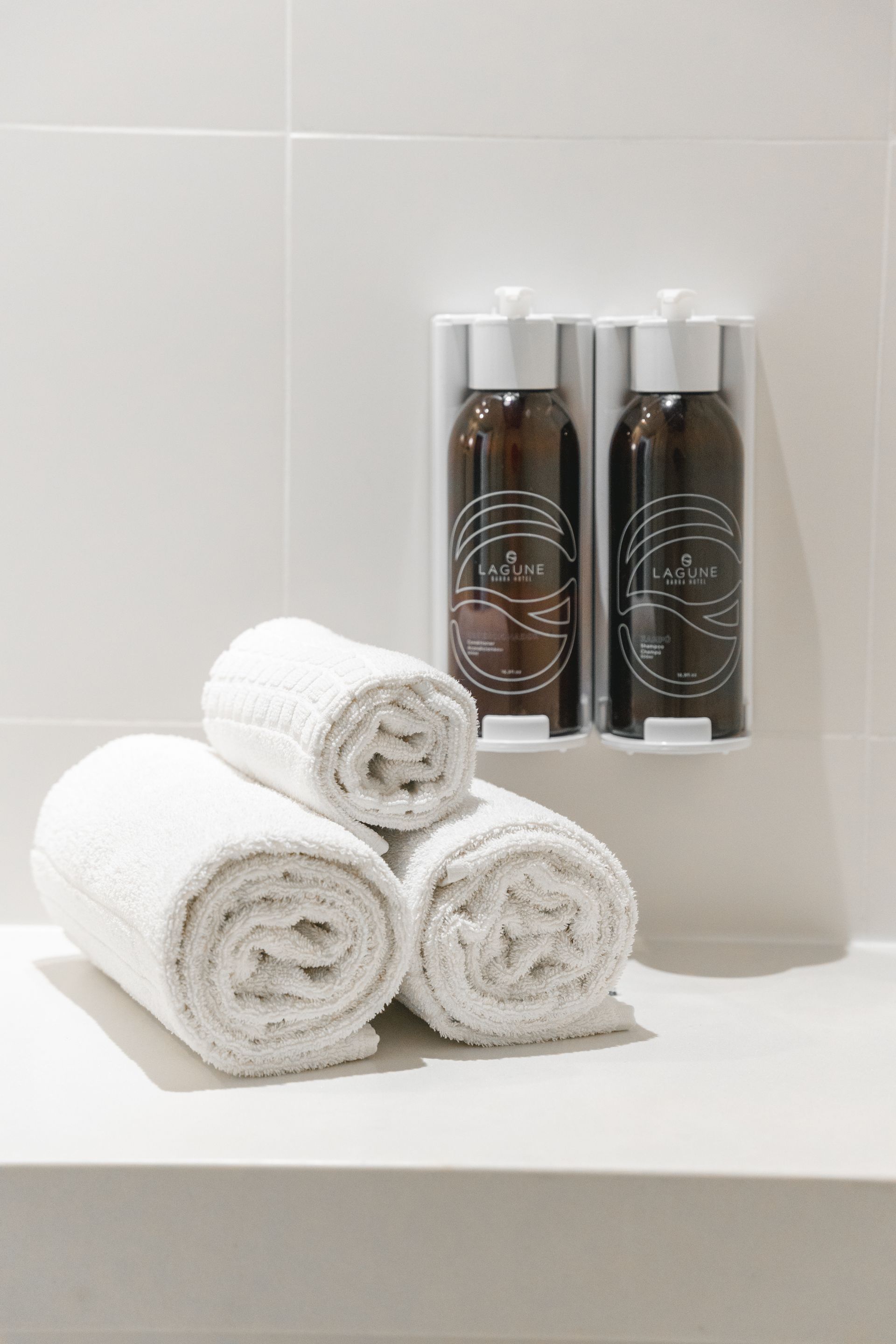 Toalhas brancas enroladas ficam em uma prateleira ao lado de dois frascos de xampu marrom no chuveiro, contra uma parede de azulejos brancos.