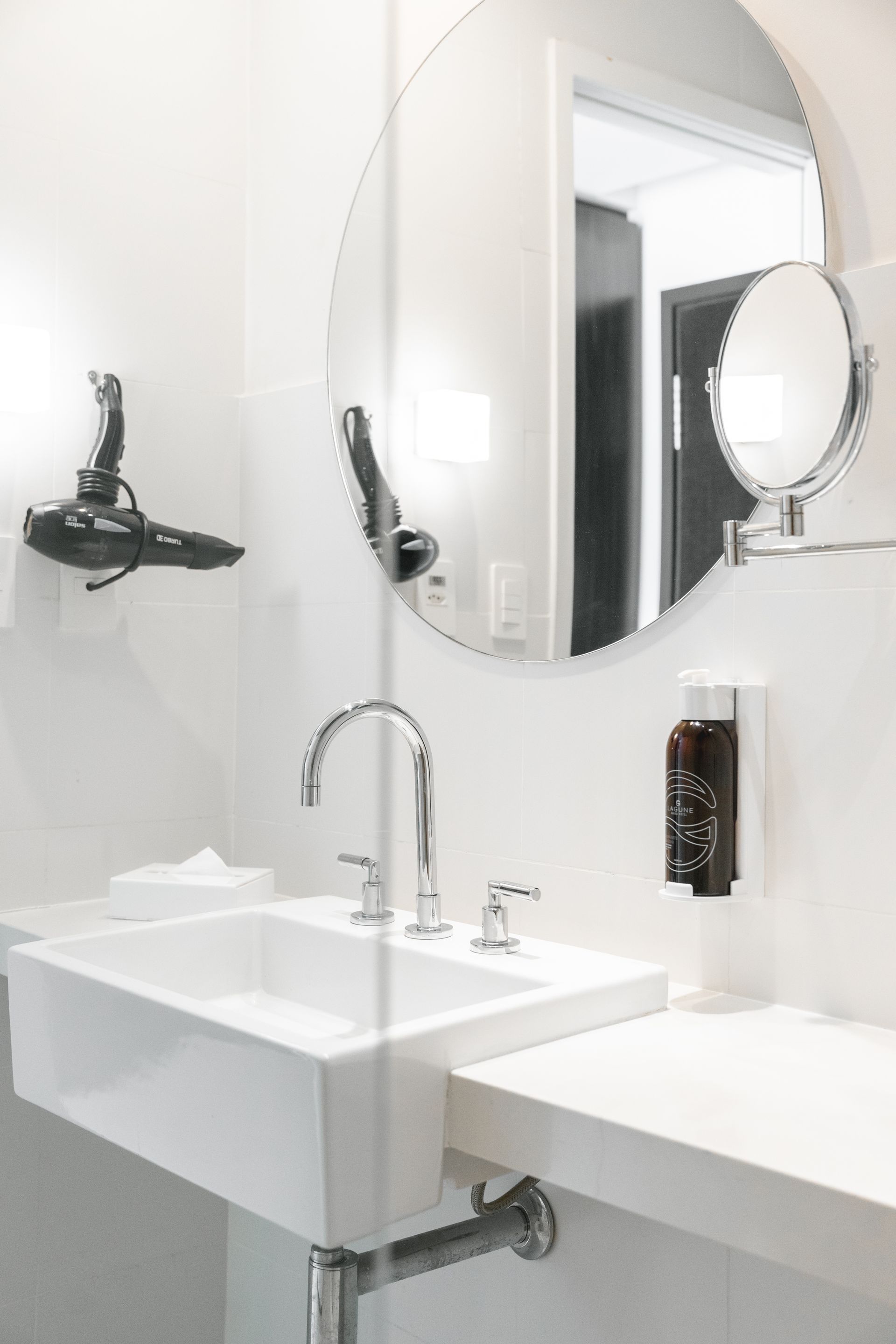 Banheiro branco com pia quadrada, espelho redondo e secador de cabelo montado.