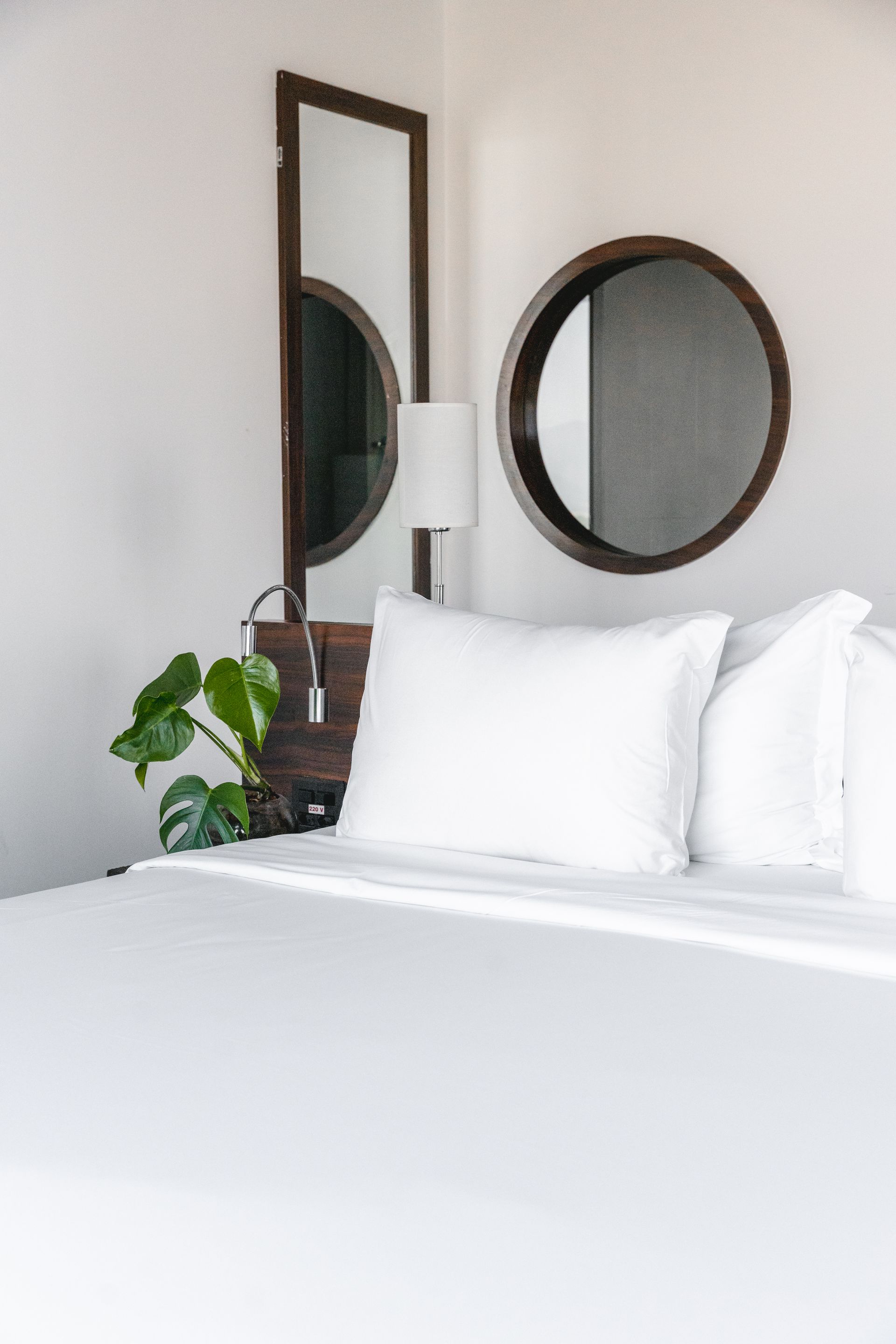 Cama branca com duas almofadas grandes, com um espelho redondo e retangular no canto e um abajur de cabeceira.