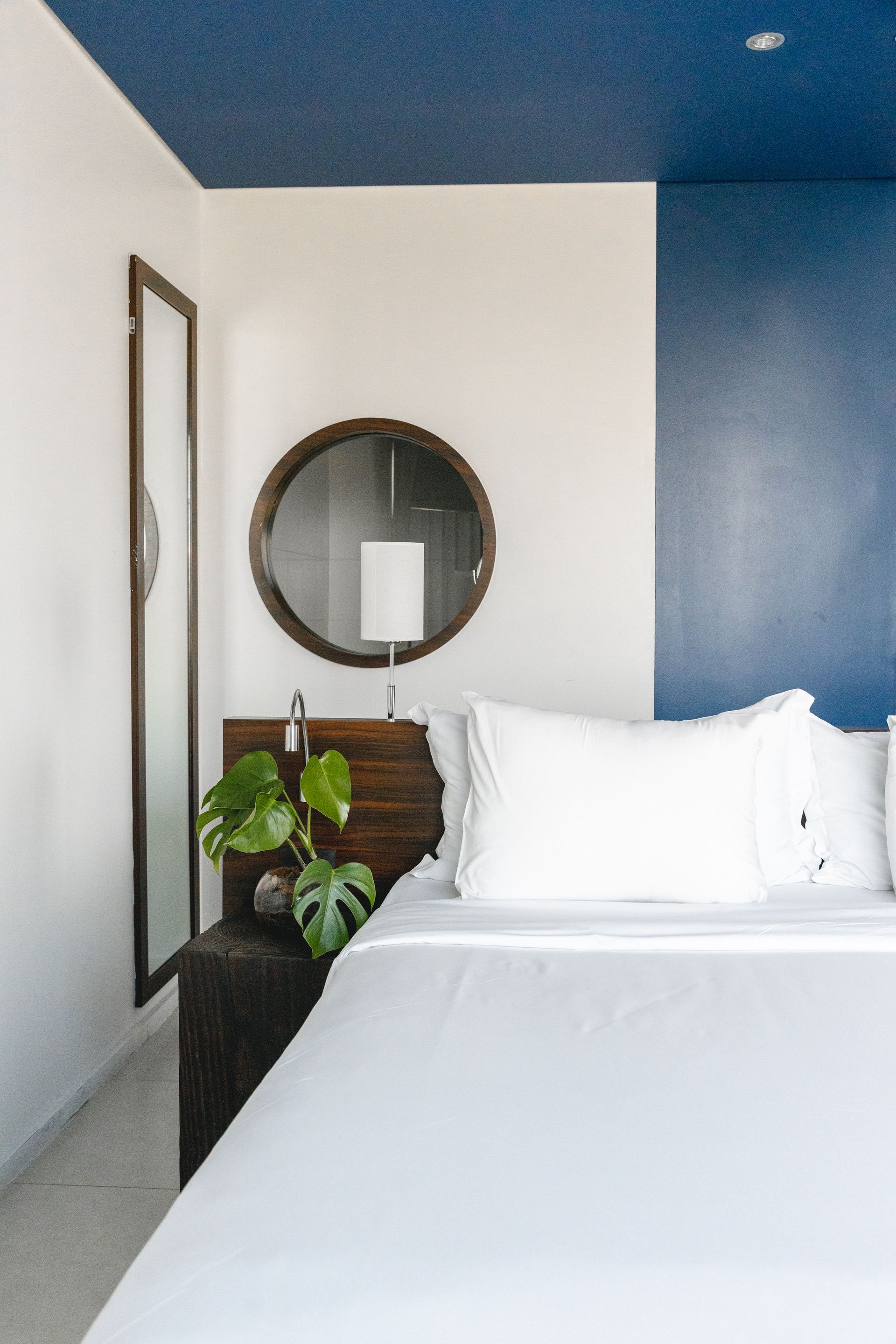 Quarto com roupa de cama branca, espelho redondo e parede com detalhes em azul-escuro. Uma planta monstera fica perto da cama.