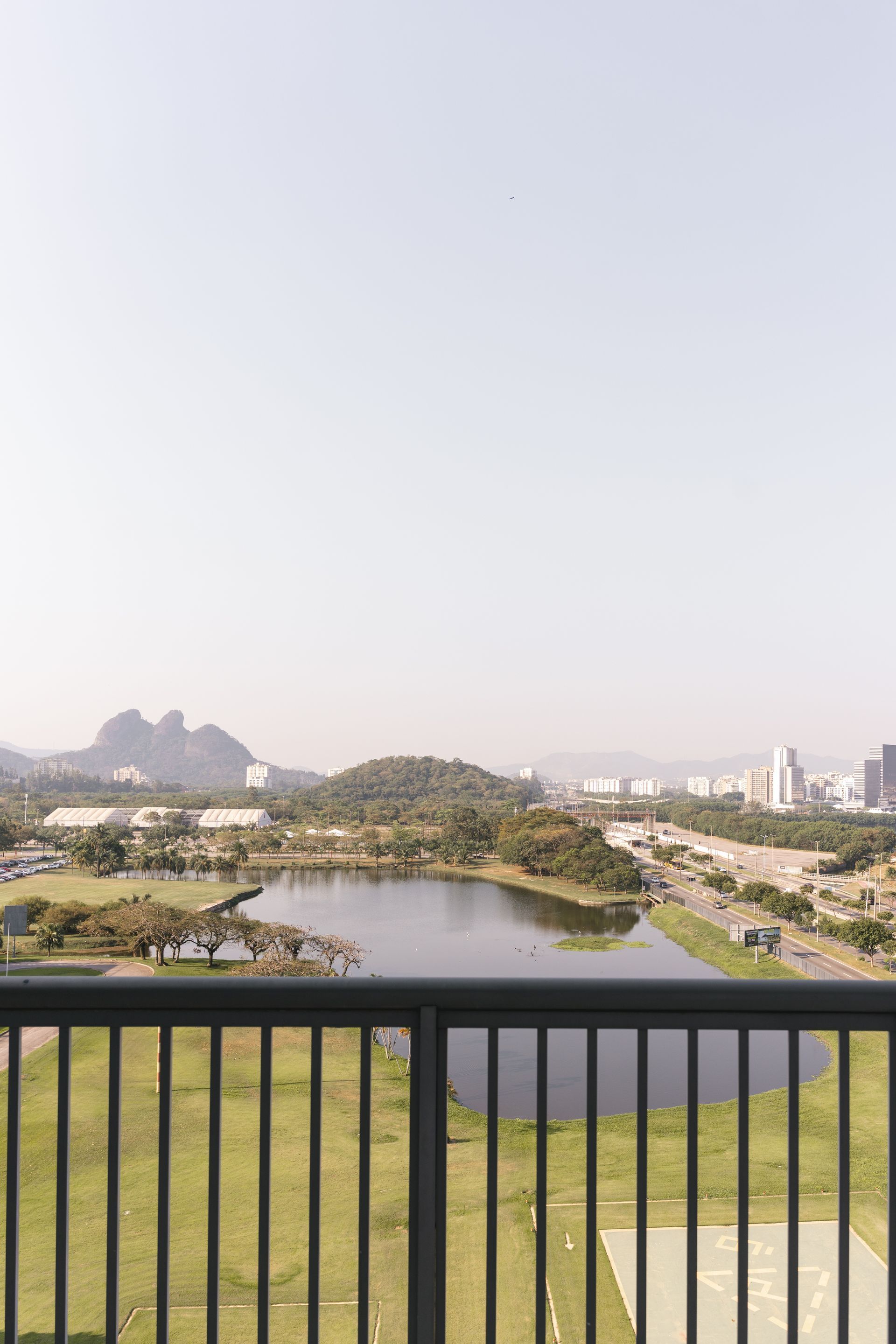 Com vista para um lago e um parque a partir de uma grade, com montanhas e uma cidade ao longe sob um céu claro.