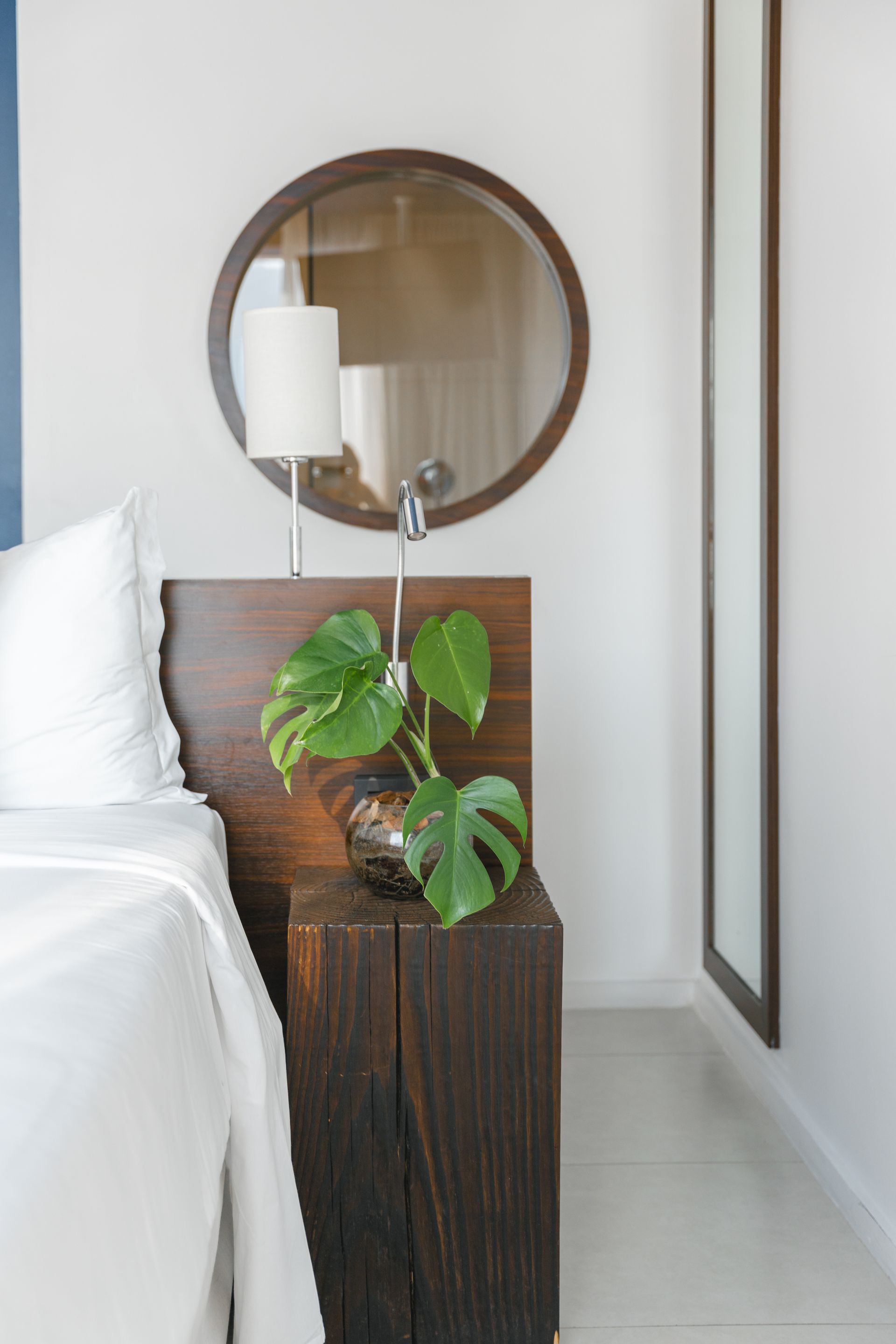 Quarto com cabeceira e mesa de cabeceira modernas de madeira escura. Uma planta Monstera repousa sobre a mesa de cabeceira. Um espelho redondo pendurado acima.