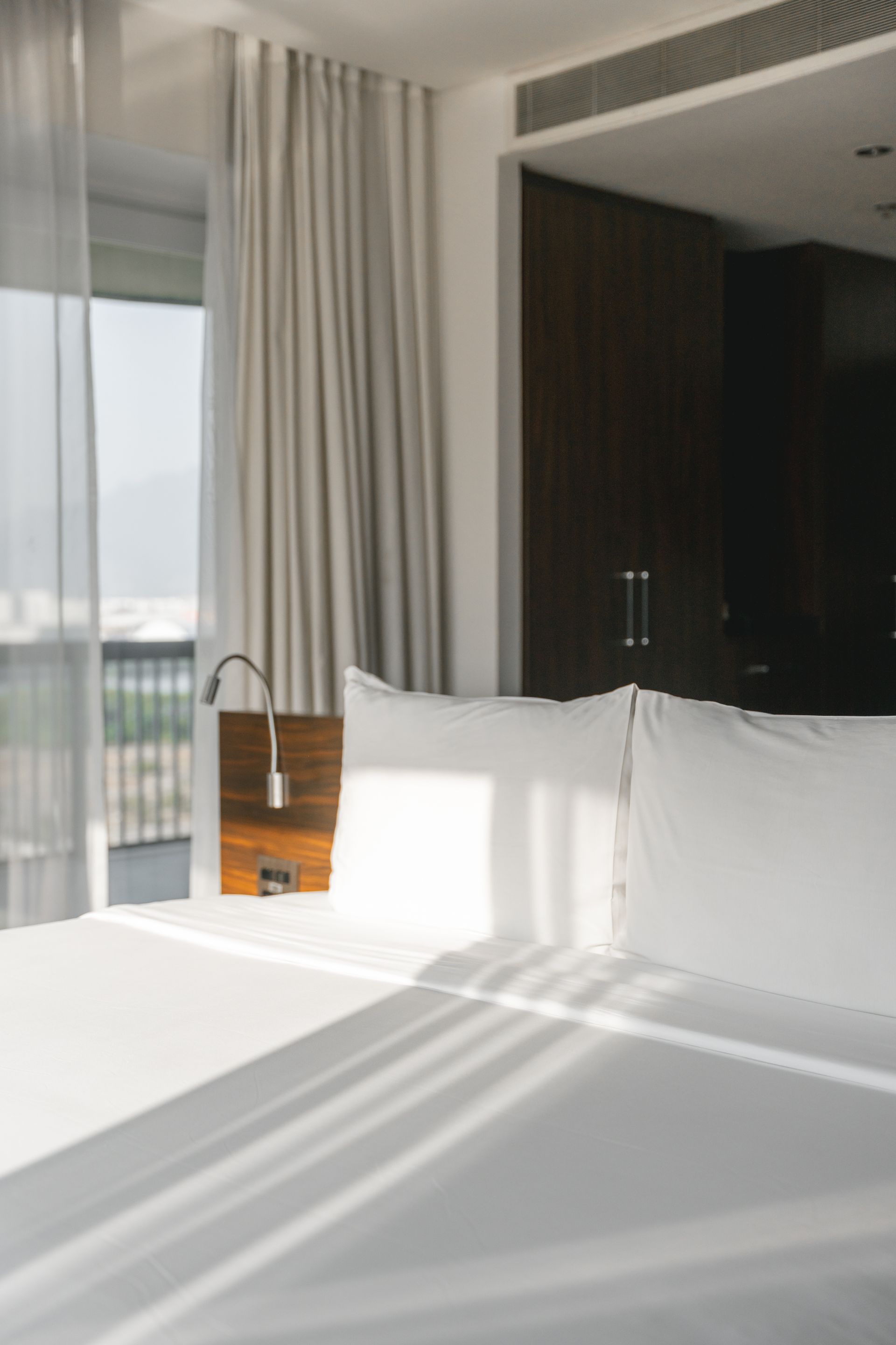 Um quarto iluminado com roupa de cama branca e luz do sol incidindo sobre a cama. Ao fundo, uma cabeceira de madeira escura e um armário.
