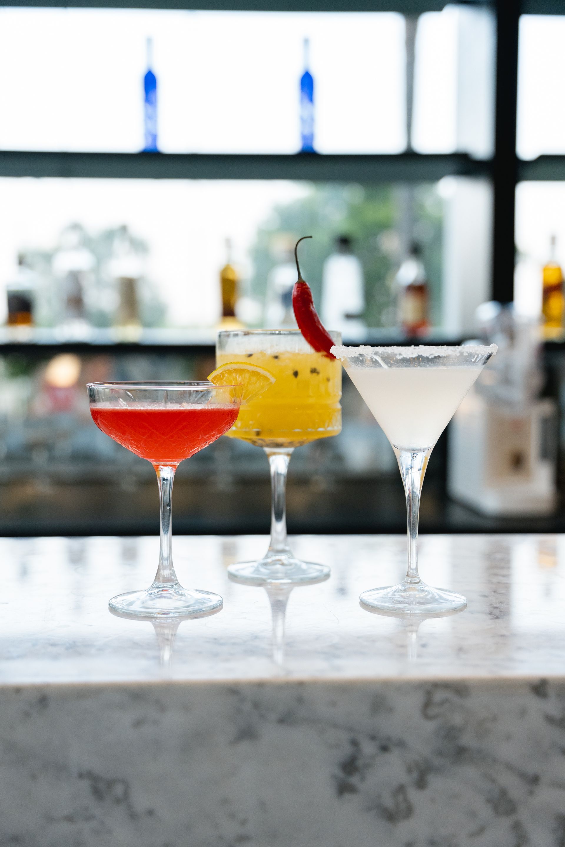 Três coquetéis estão dispostos em um balcão: um tinto, um amarelo com pimenta e um branco com borda açucarada. O balcão fica em frente a uma prateleira com garrafas.