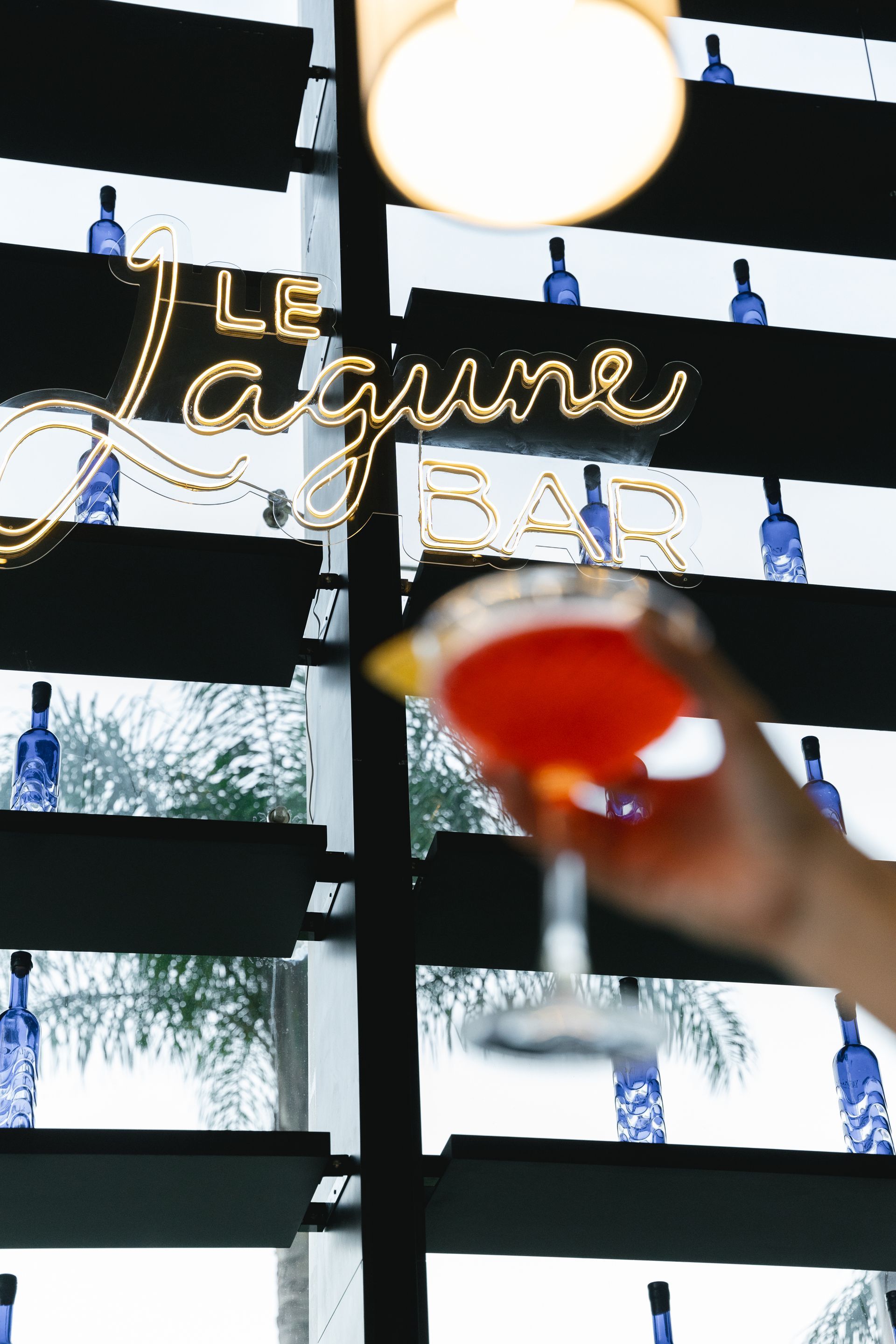 Uma pessoa segura um coquetel no Le Lagune Bar. O letreiro de neon brilha, com uma garrafa azul ao fundo.