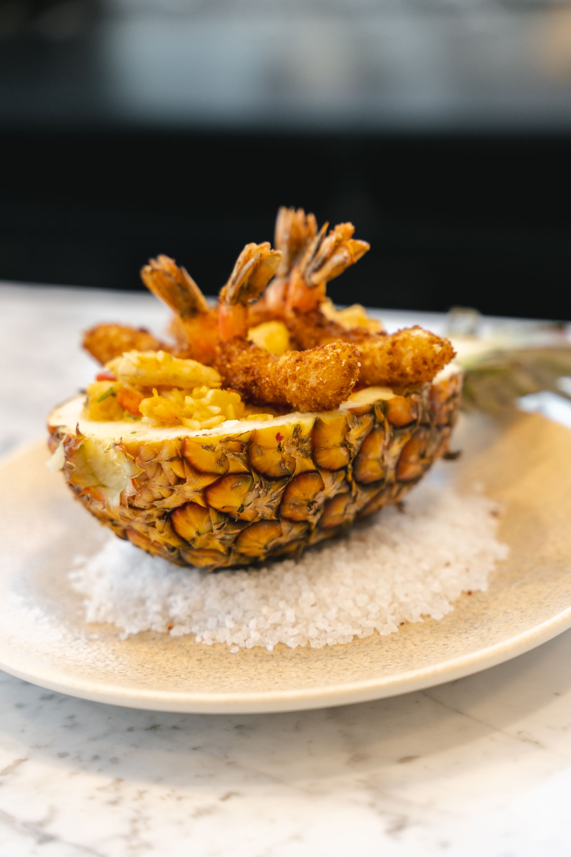 Camarão frito e pedaços de abacaxi servidos em uma metade de abacaxi oca, sobre uma cama de sal em um prato.