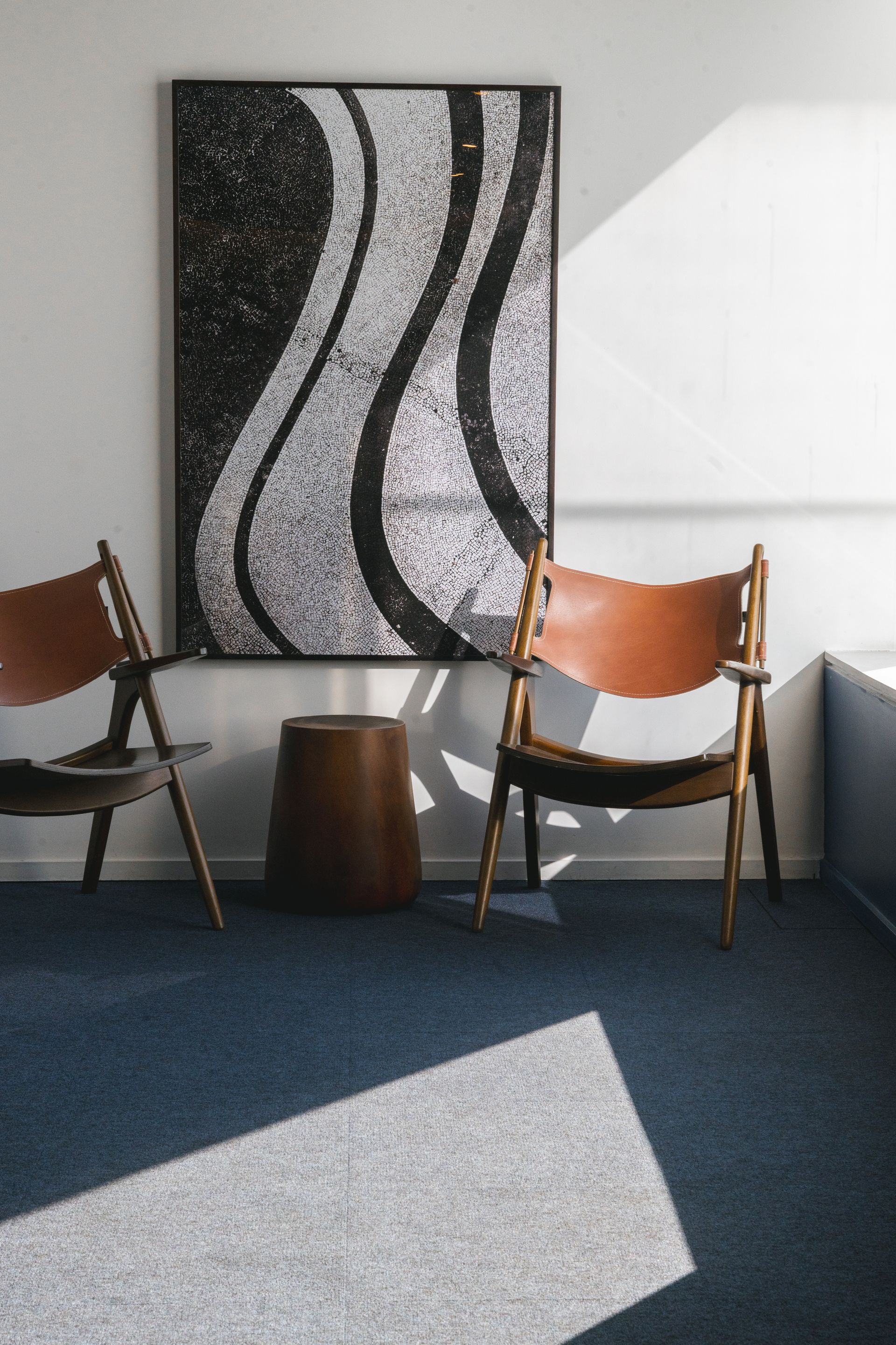 Duas cadeiras de meados do século com assentos de couro marrom ladeiam uma mesa marrom em formato de tambor. Uma obra de arte em mosaico preto e branco está pendurada na parede.