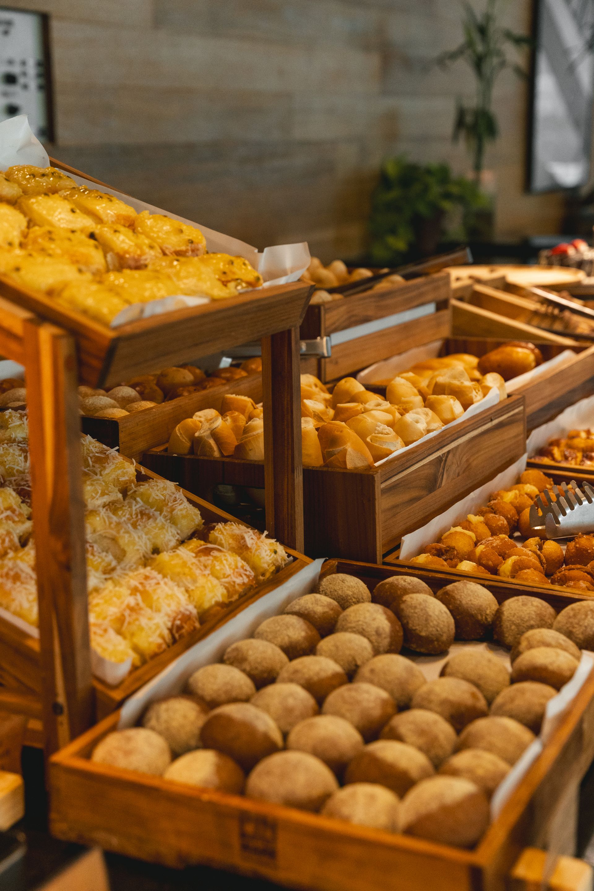 Expositor de madeira repleto de diversos produtos assados, incluindo pãezinhos e doces. Ambiente de bufê.
