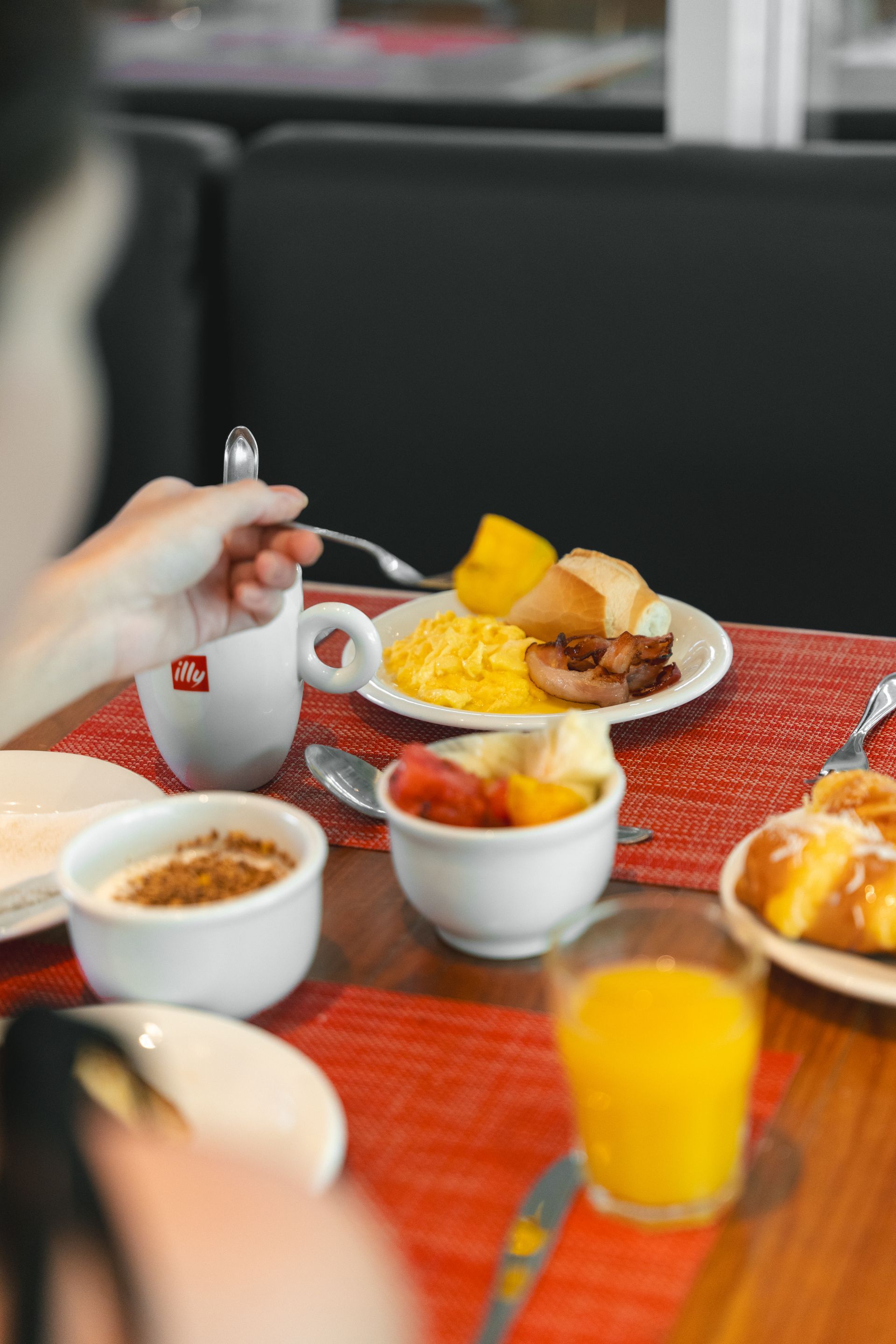 Pessoa tomando café da manhã em uma mesa com ovos mexidos, frutas, torradas, café, suco e doces.