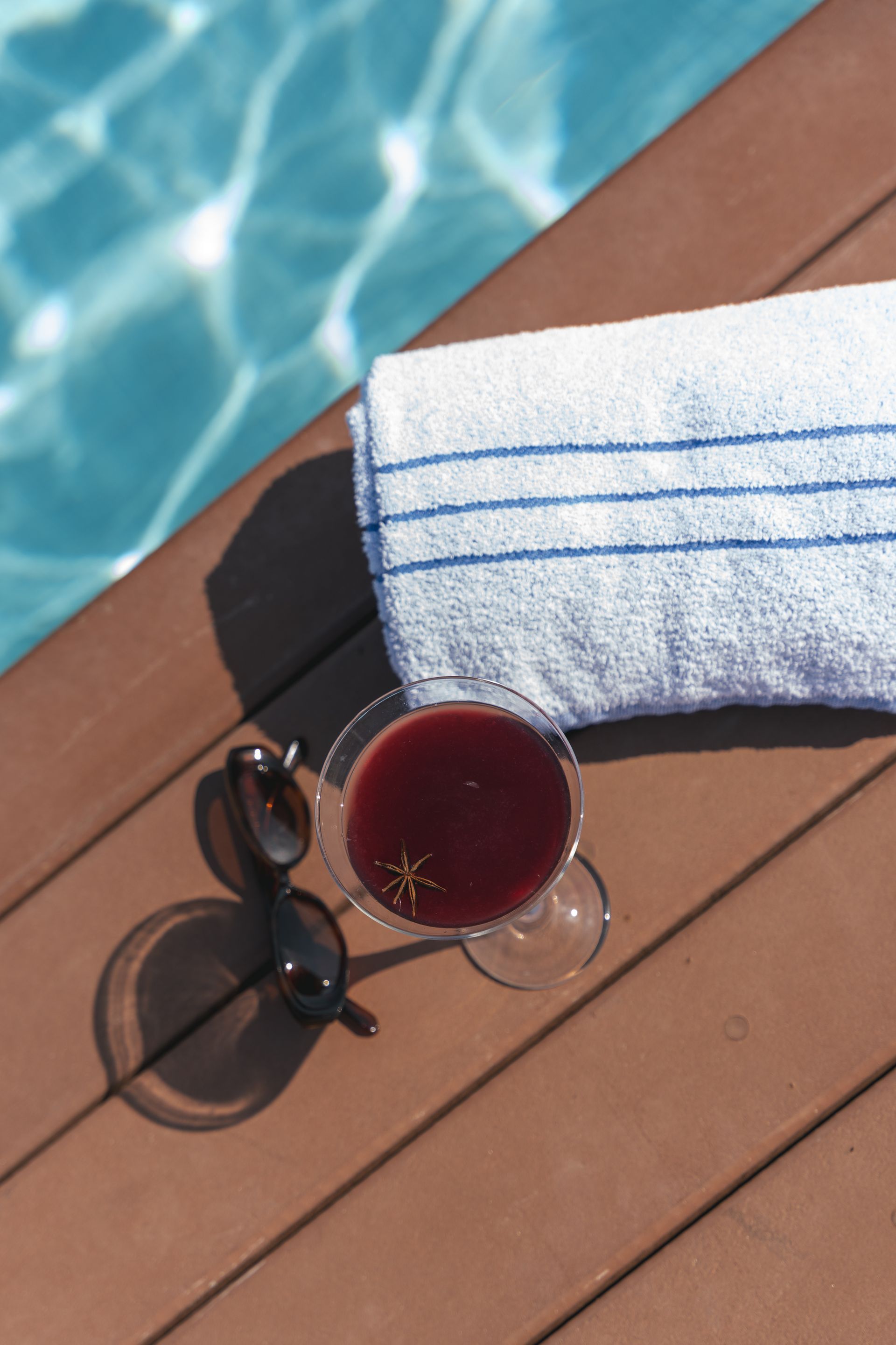 Um coquetel e óculos de sol ao lado de uma toalha dobrada estão em um deck marrom, com uma piscina azul ao fundo.