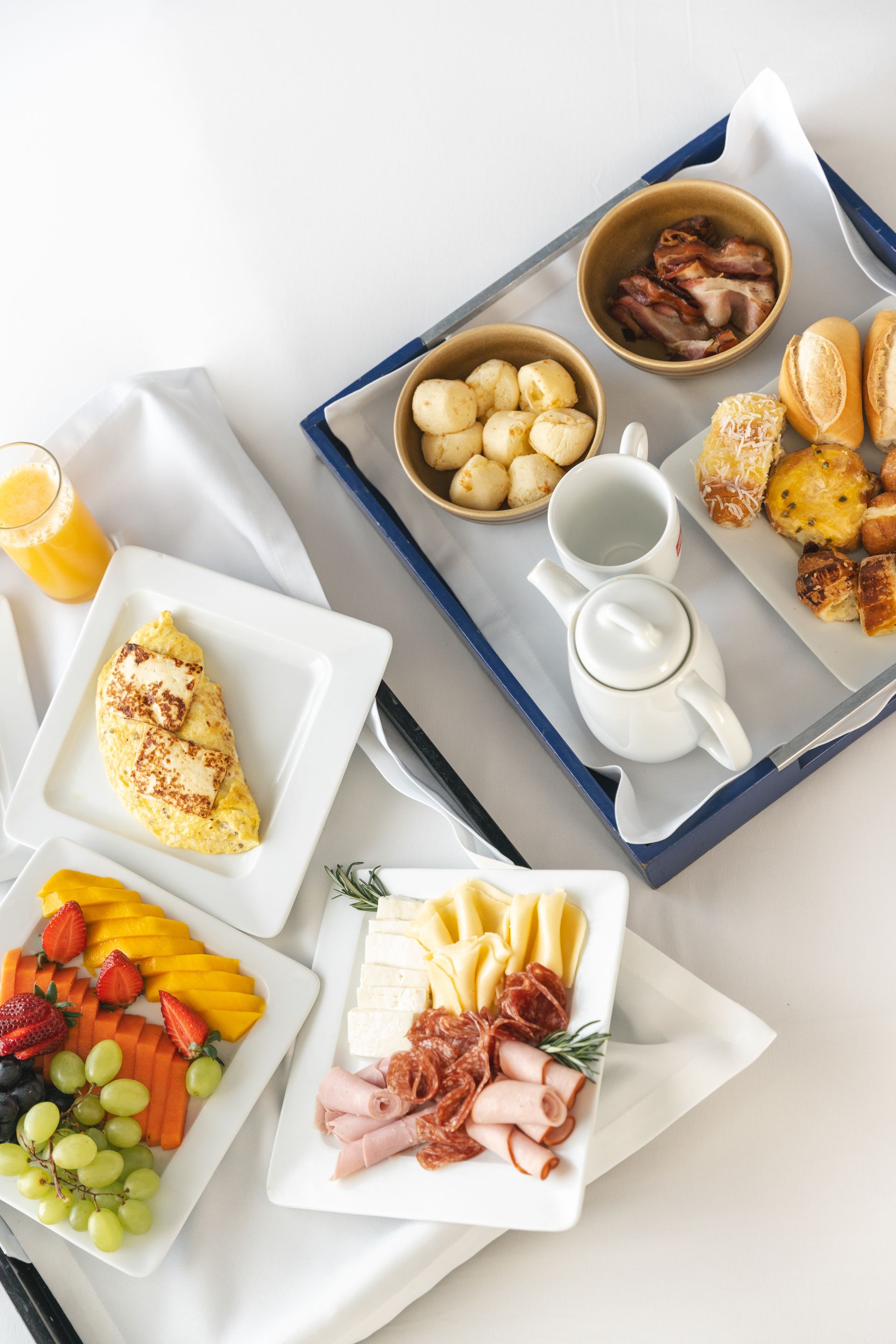 Café da manhã com frutas, ovos, pão, carne e suco em uma mesa branca com uma bandeja azul.