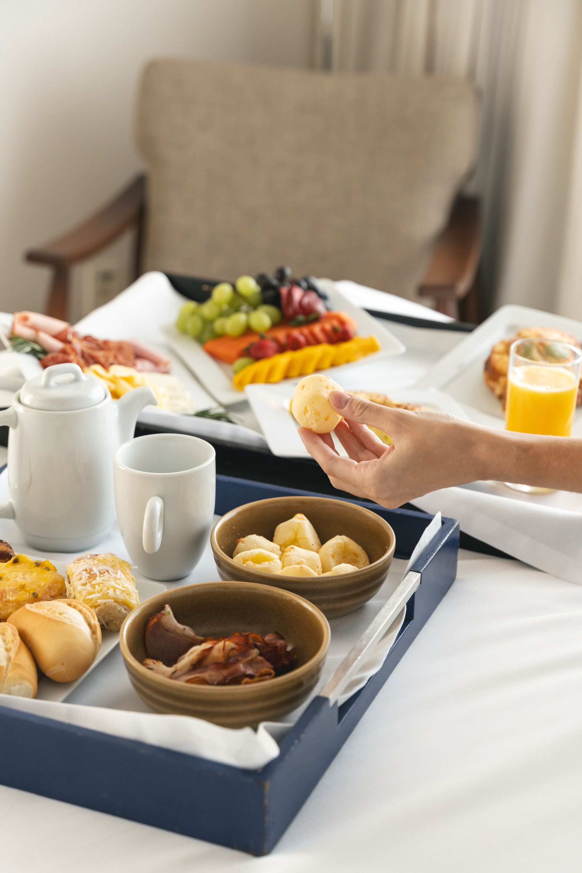 Bandeja de café da manhã sobre uma cama: frutas, doces, suco e uma mão estendendo a mão para pegar comida. Uma cadeira em tom neutro ao fundo.