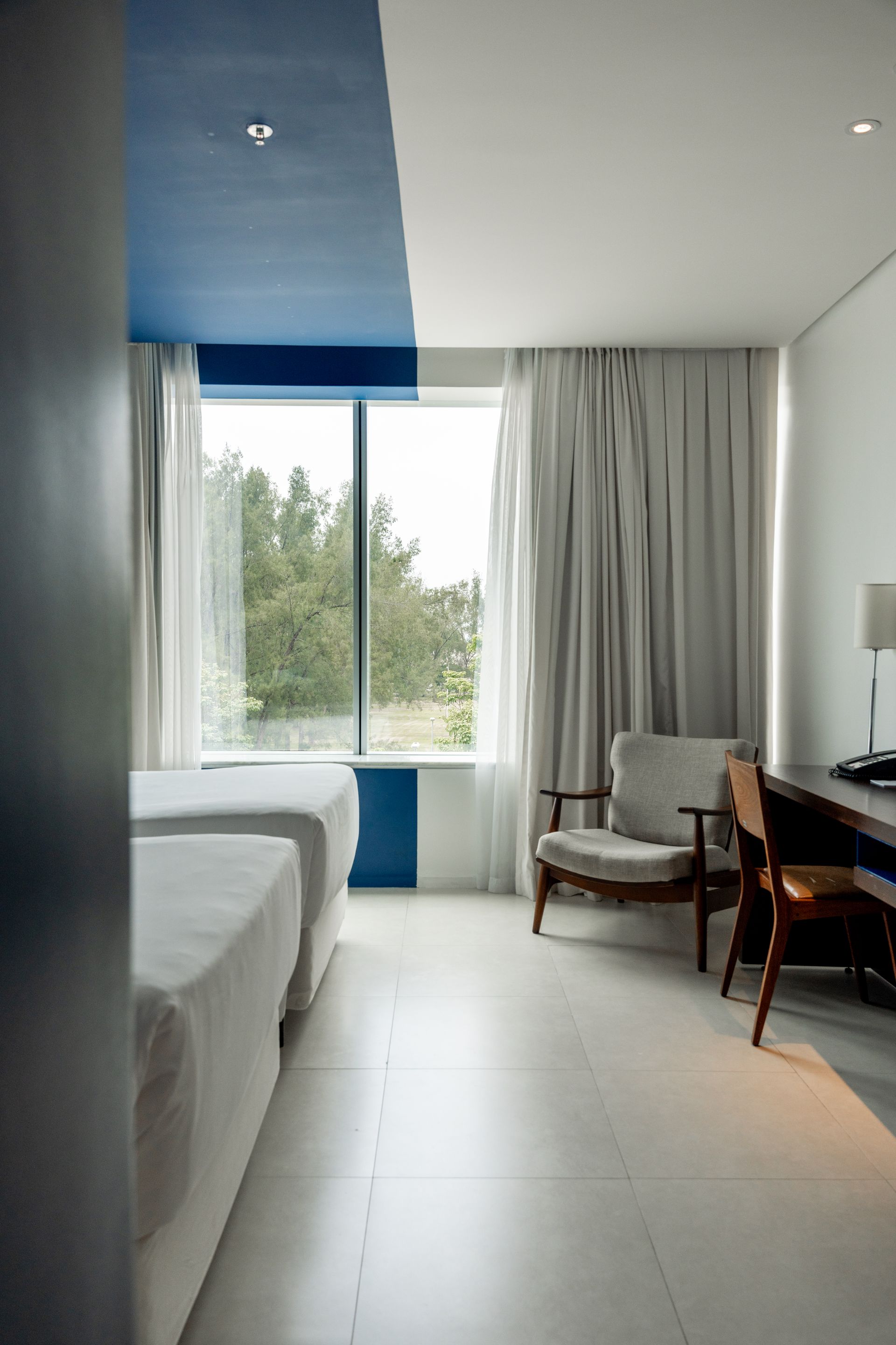Um quarto de hotel com duas camas, uma cadeira, escrivaninha e uma janela grande. O teto é azul e branco, com paredes brancas.