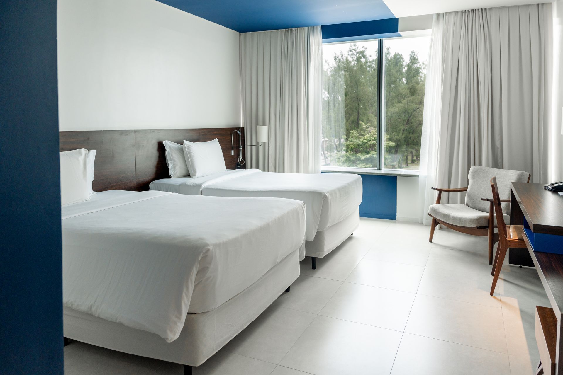 Um quarto de hotel bem iluminado com duas camas de solteiro, uma escrivaninha e uma cadeira. Possui paredes brancas, detalhes em azul e uma grande janela com vista para as árvores.
