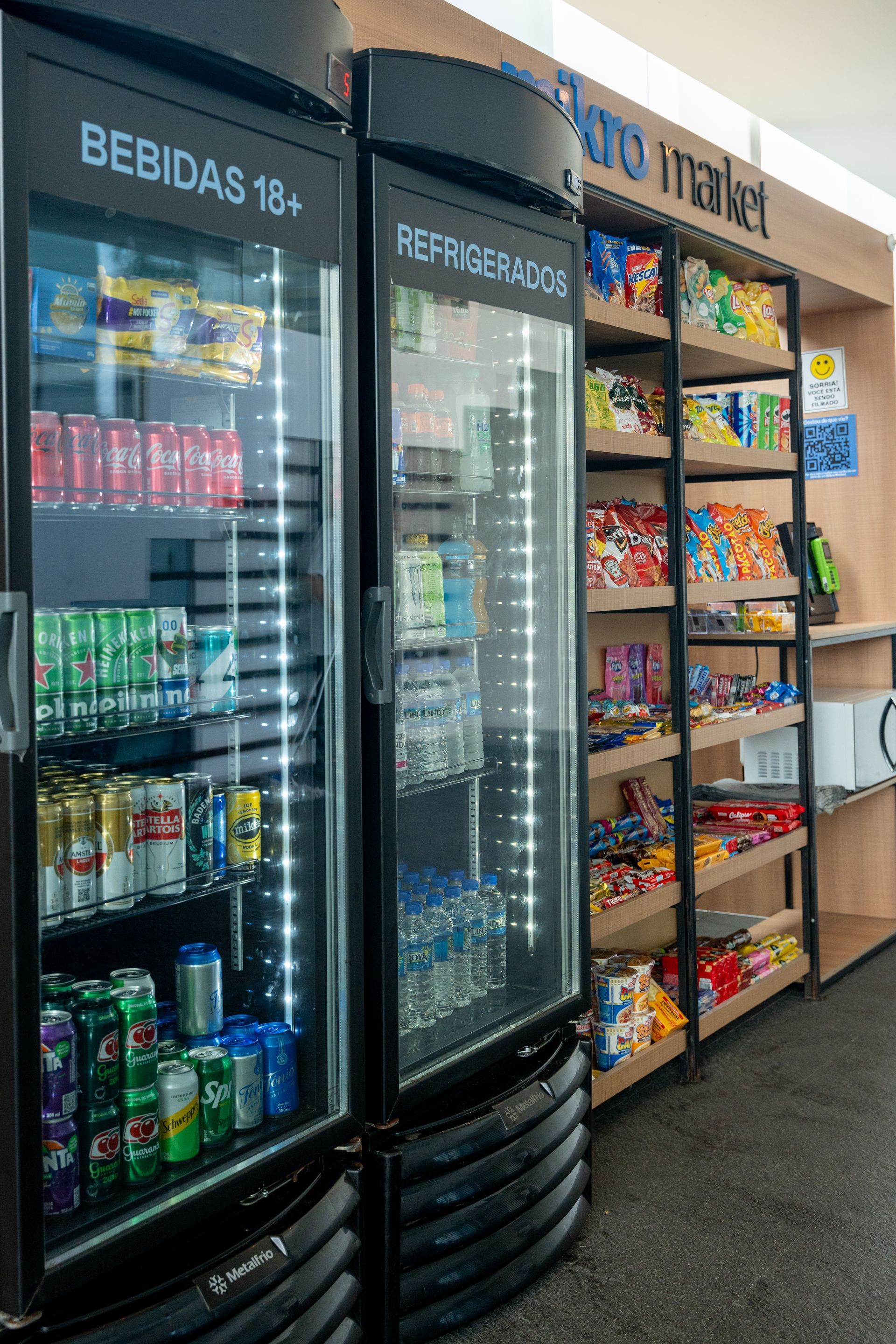 Um pequeno mercado com geladeiras abastecidas com bebidas e uma prateleira com salgadinhos. As bebidas estão em latas vermelhas, verdes e azuis.