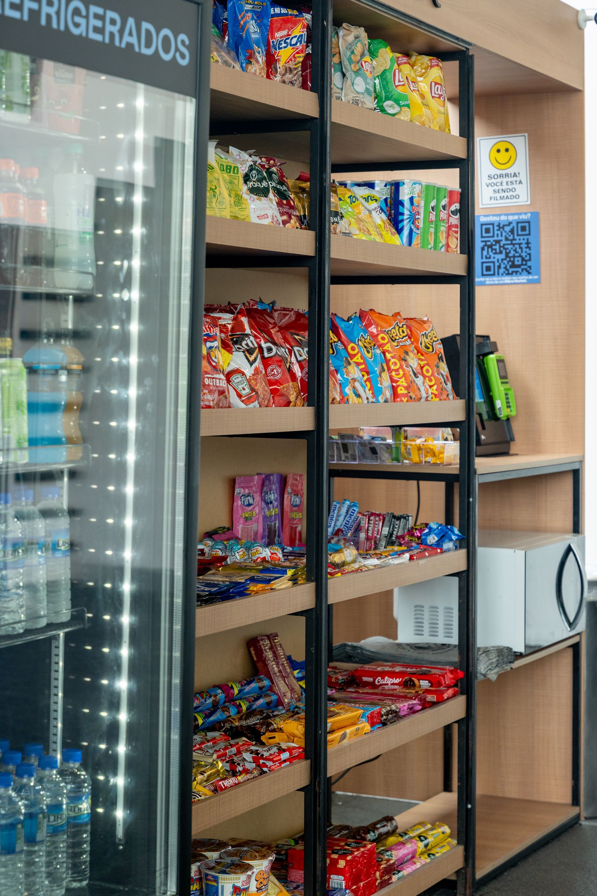 Uma lanchonete com prateleiras de salgadinhos, doces e uma geladeira com bebidas. Há um micro-ondas em um balcão próximo.