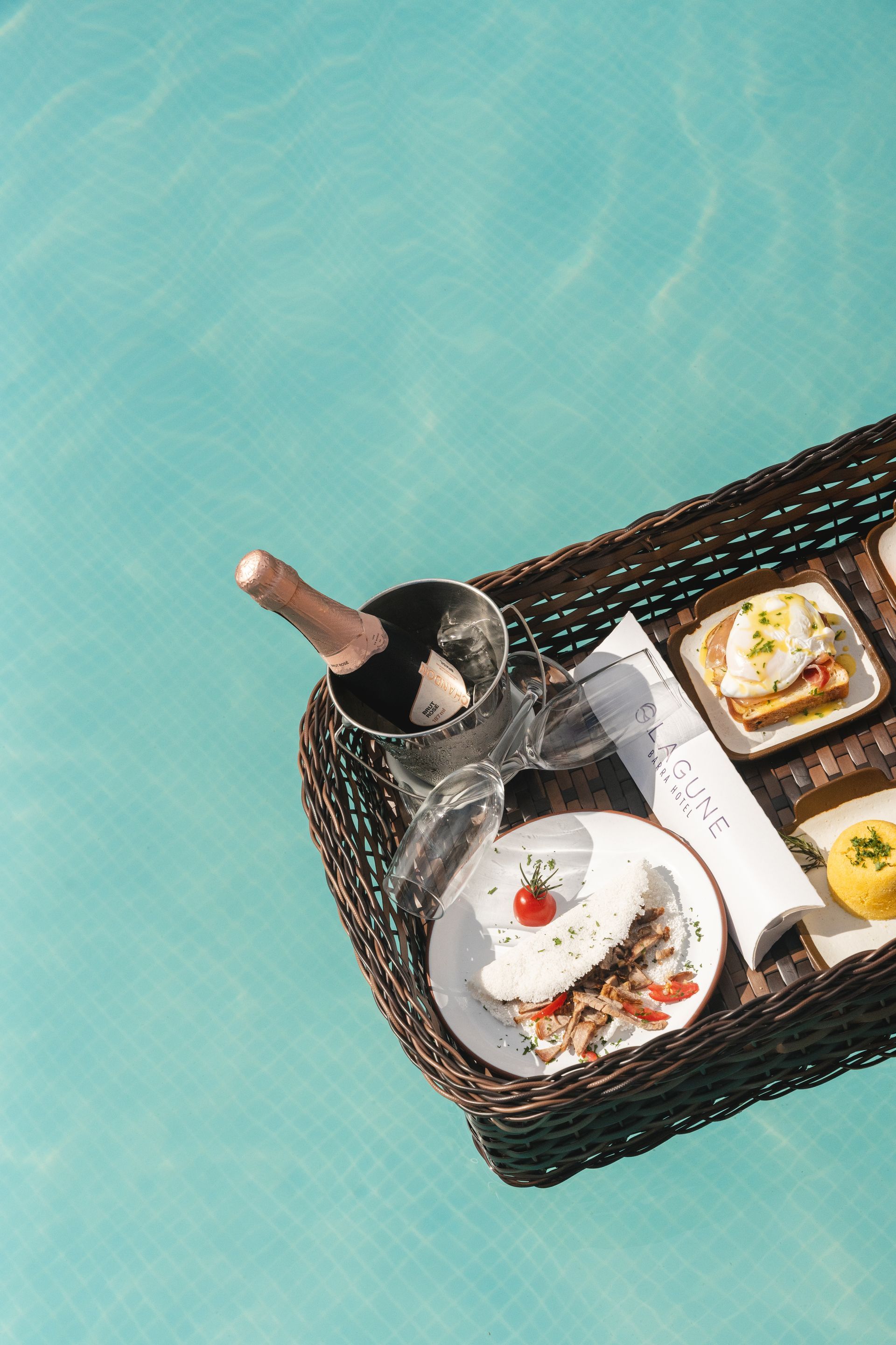 Bandeja de café da manhã com champanhe e comida flutua em uma piscina azul brilhante.