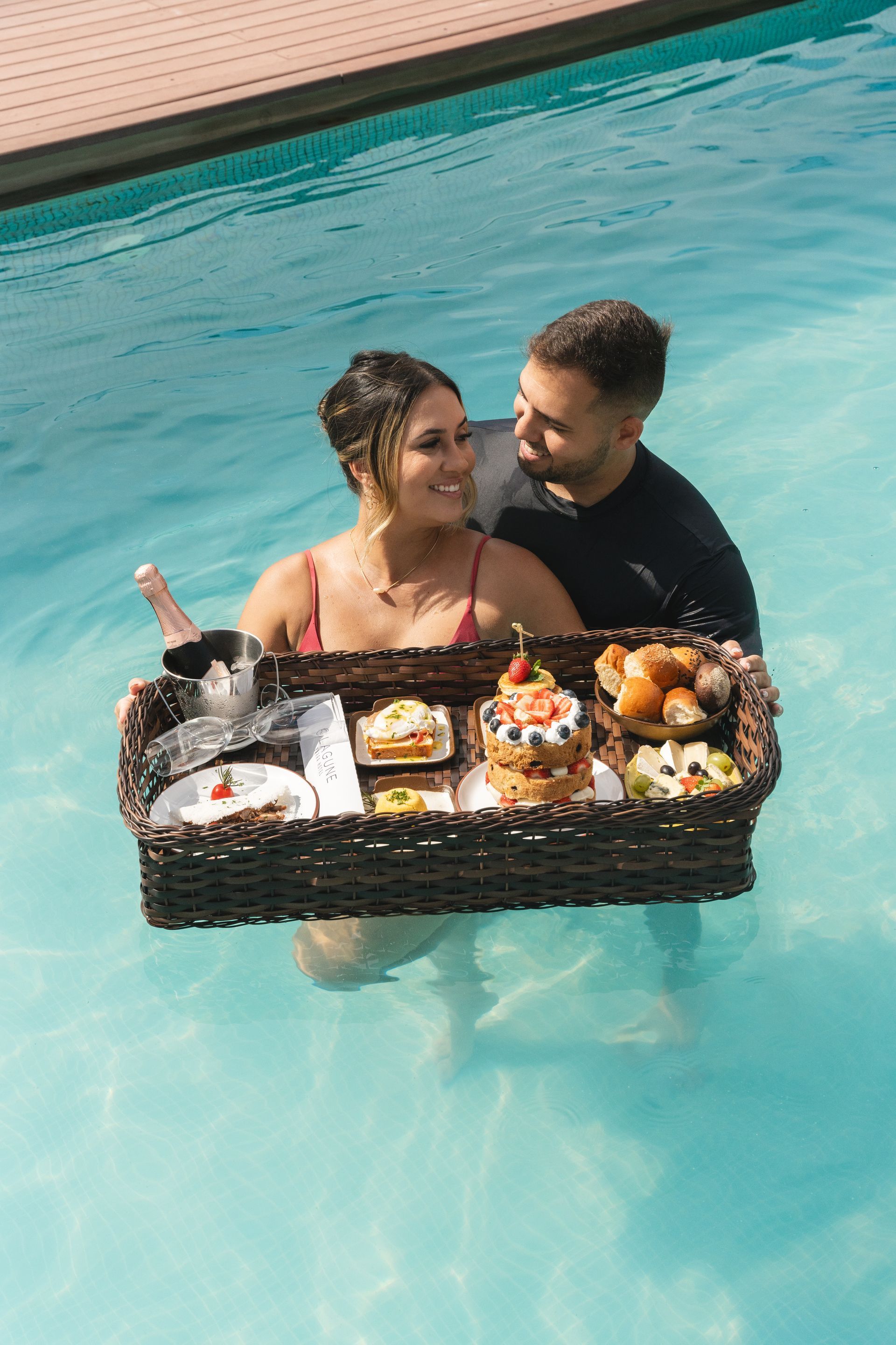 Casal tomando café da manhã na piscina. Mulher sorri para homem, que segura uma bandeja com comida e champanhe.
