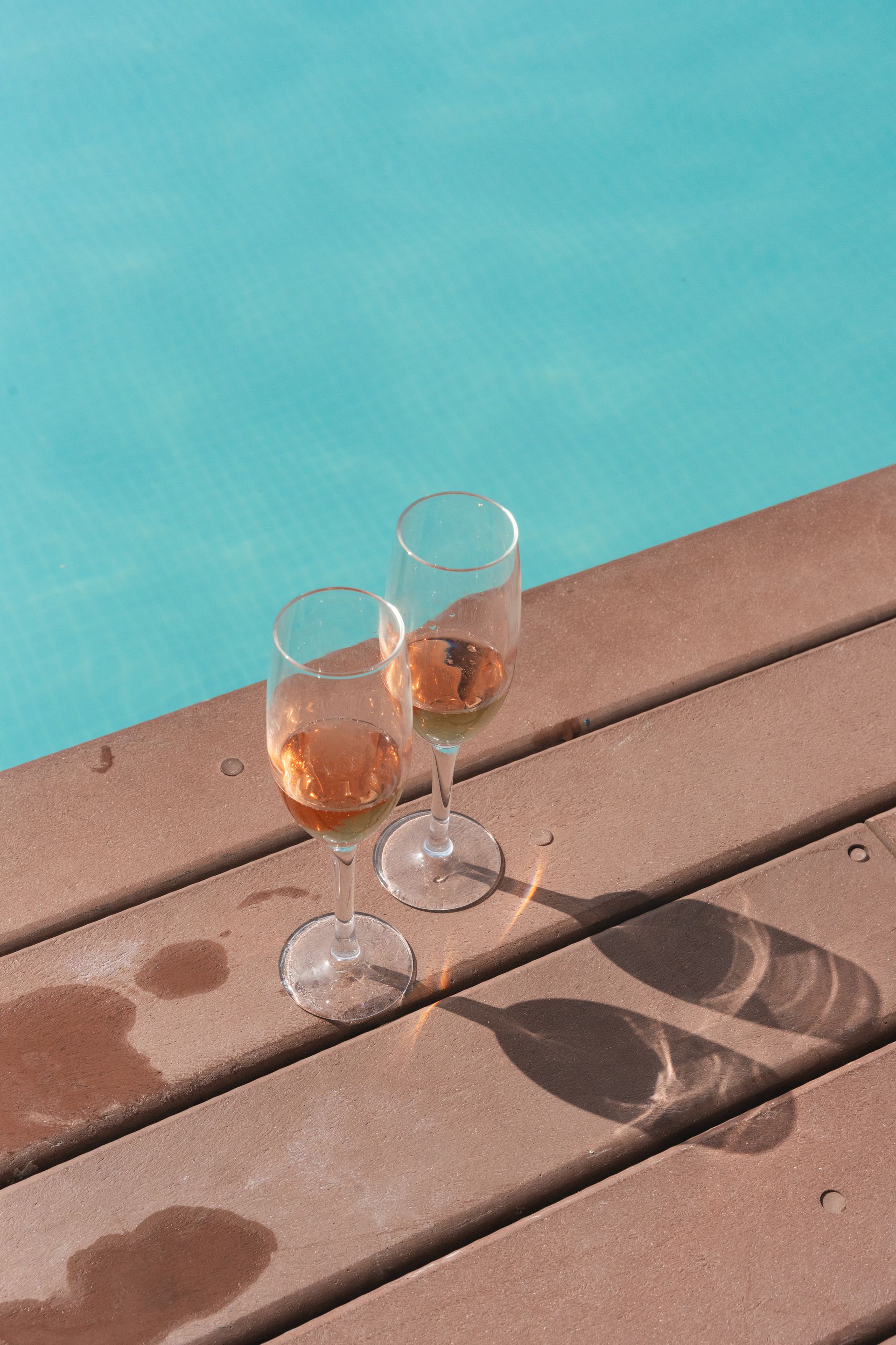 Duas taças de vinho rosé em um deck de madeira ao lado de uma piscina azul-turquesa.