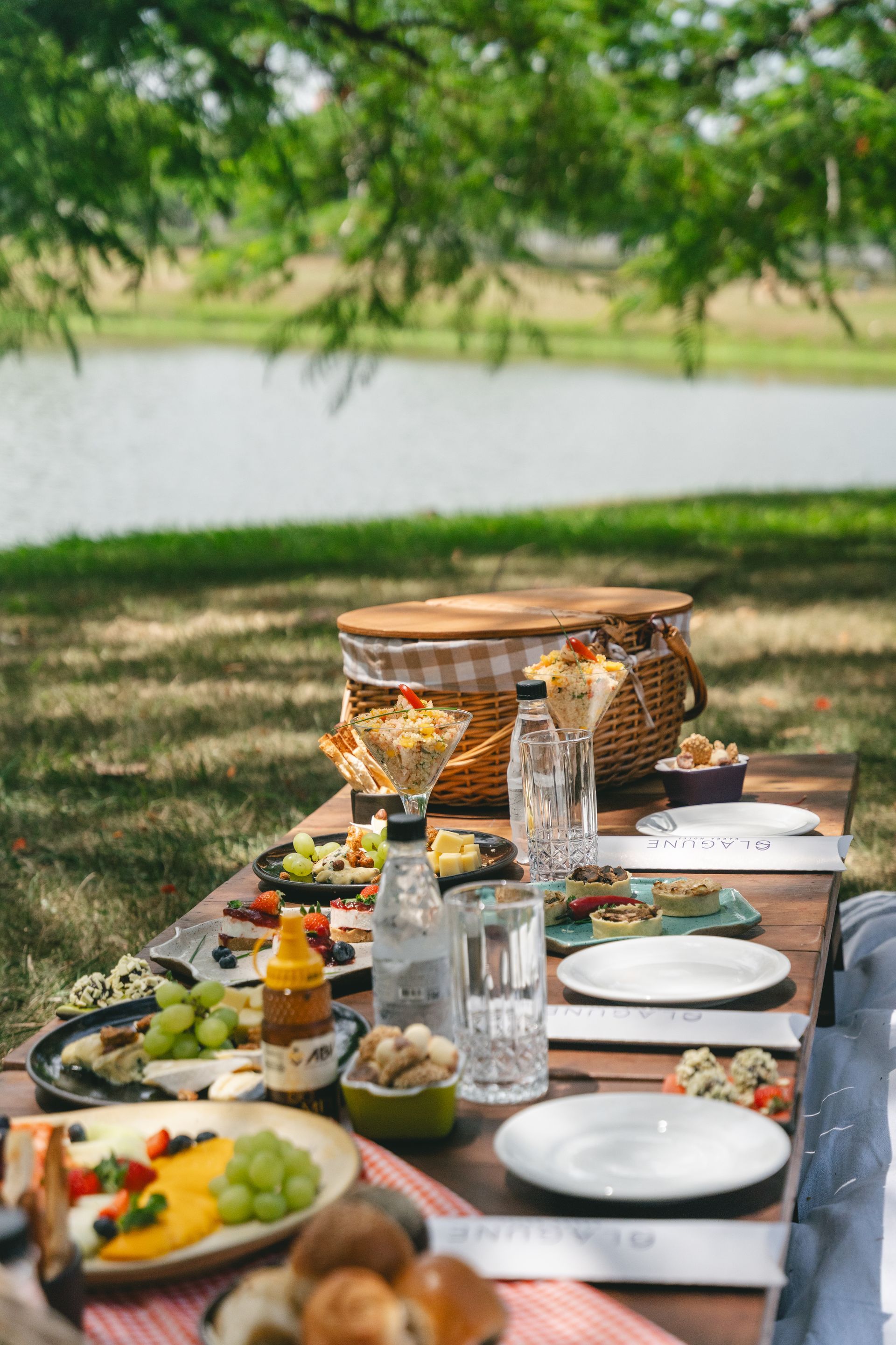 Mesa de piquenique com comida e bebidas em frente a um lago; uma cesta de vime ao fundo.