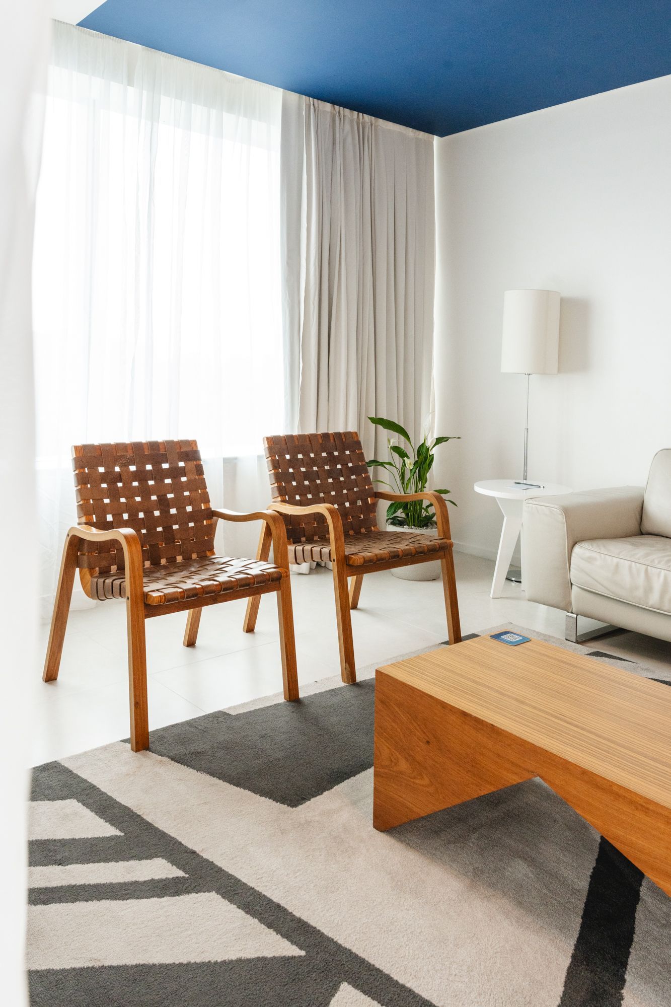 Duas poltronas de couro trançado estão de frente para uma mesa de centro em uma sala de estar moderna com teto azul e paredes brancas. Um tapete estampado e cortinas transparentes também são visíveis.