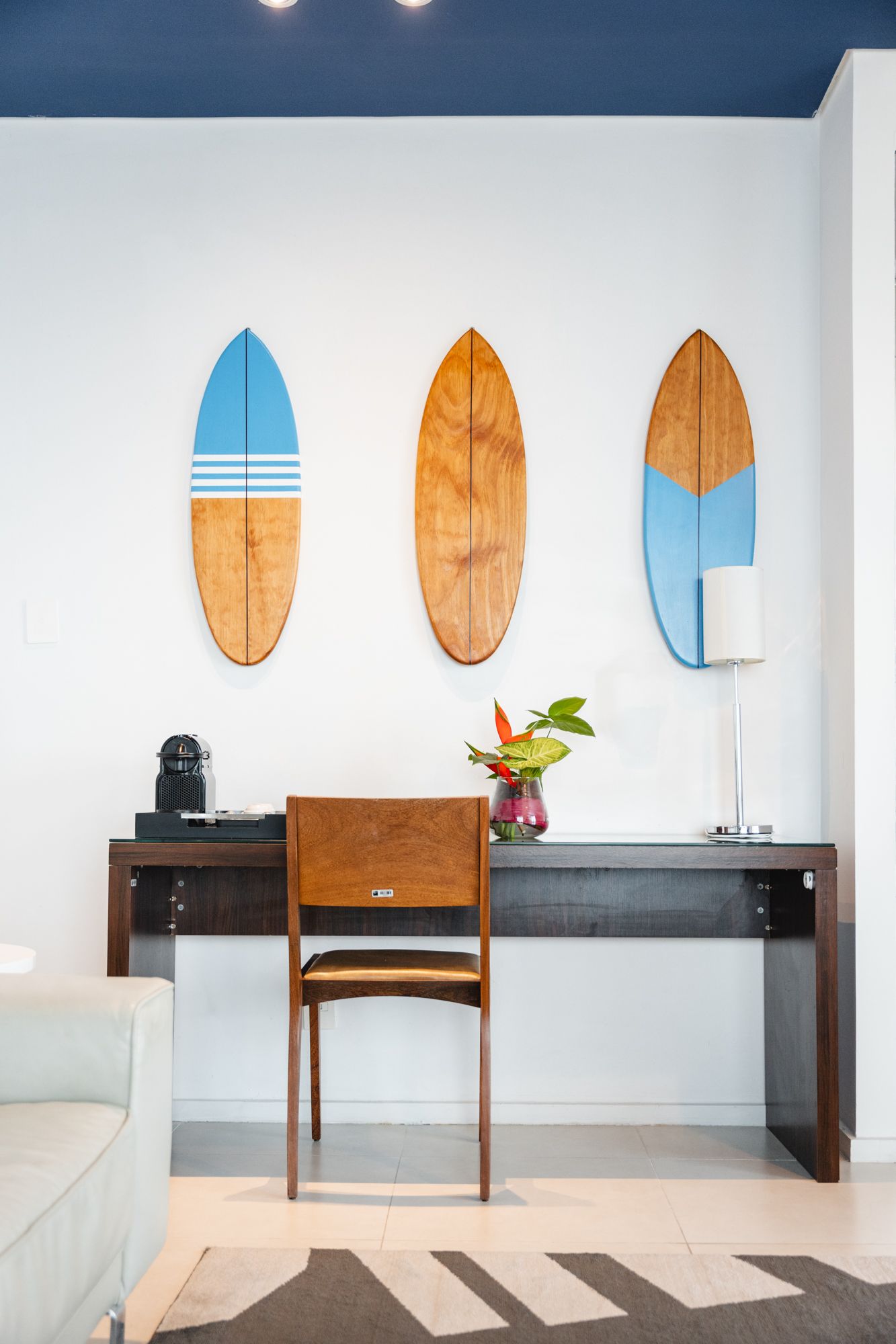 Três pranchas de surfe penduradas sobre uma mesa com uma cadeira. Um abajur e um vaso de flores repousam sobre a mesa.