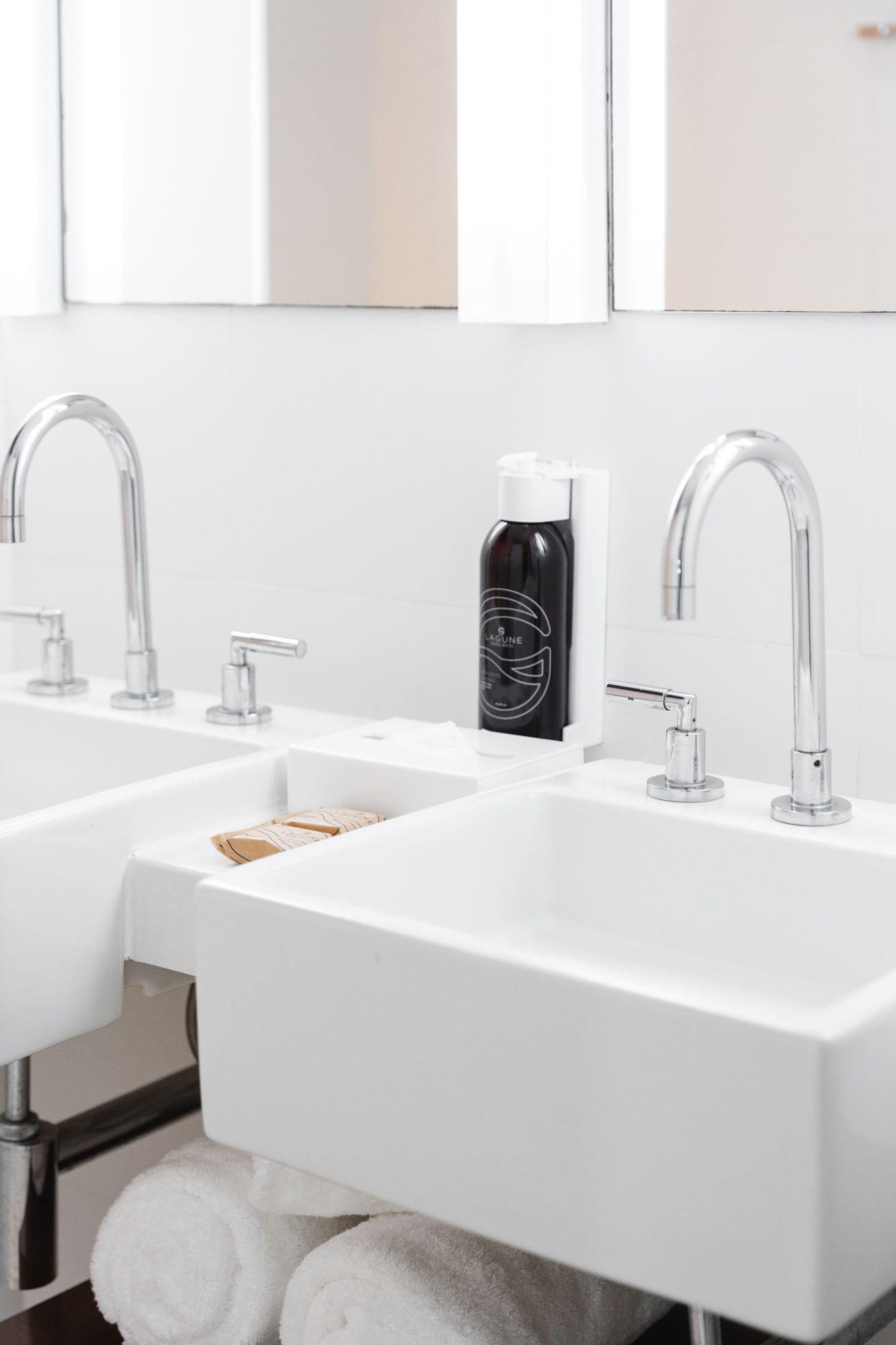 Duas pias brancas com torneiras cromadas em um banheiro moderno. Entre elas, há um frasco de sabonete e toalhas empilhadas abaixo.