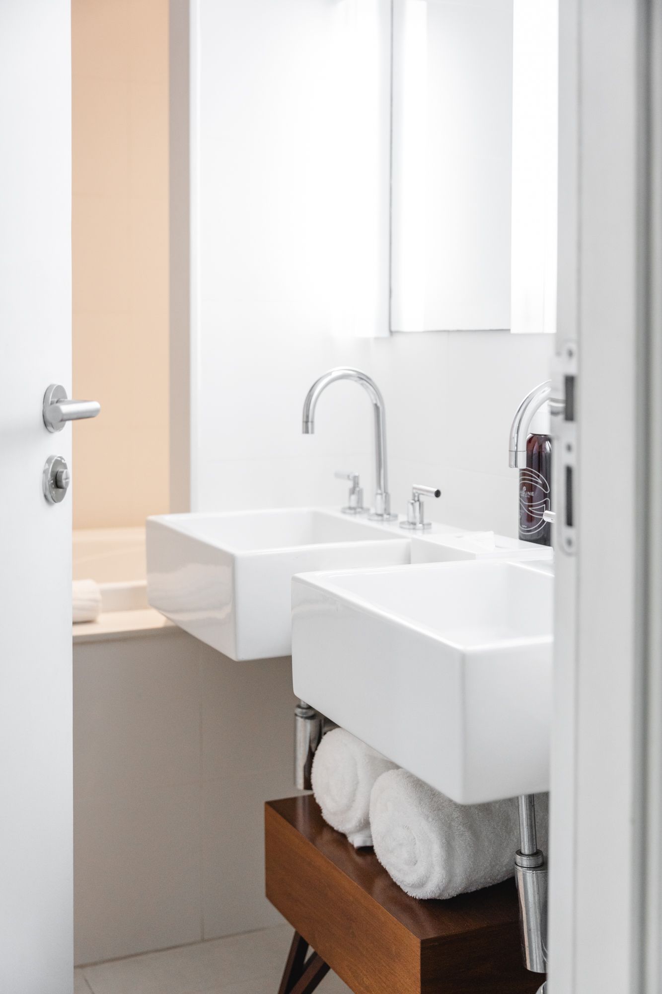 Banheiro moderno e iluminado com duas pias brancas quadradas sobre um armário de madeira escura. Toalhas brancas enroladas empilhadas abaixo das pias.