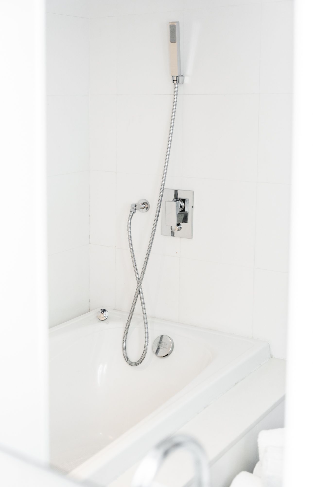 Banheira branca com chuveiro de mão e torneira encostada em uma parede de azulejos brancos. Uma corrente de metal segura o chuveiro.