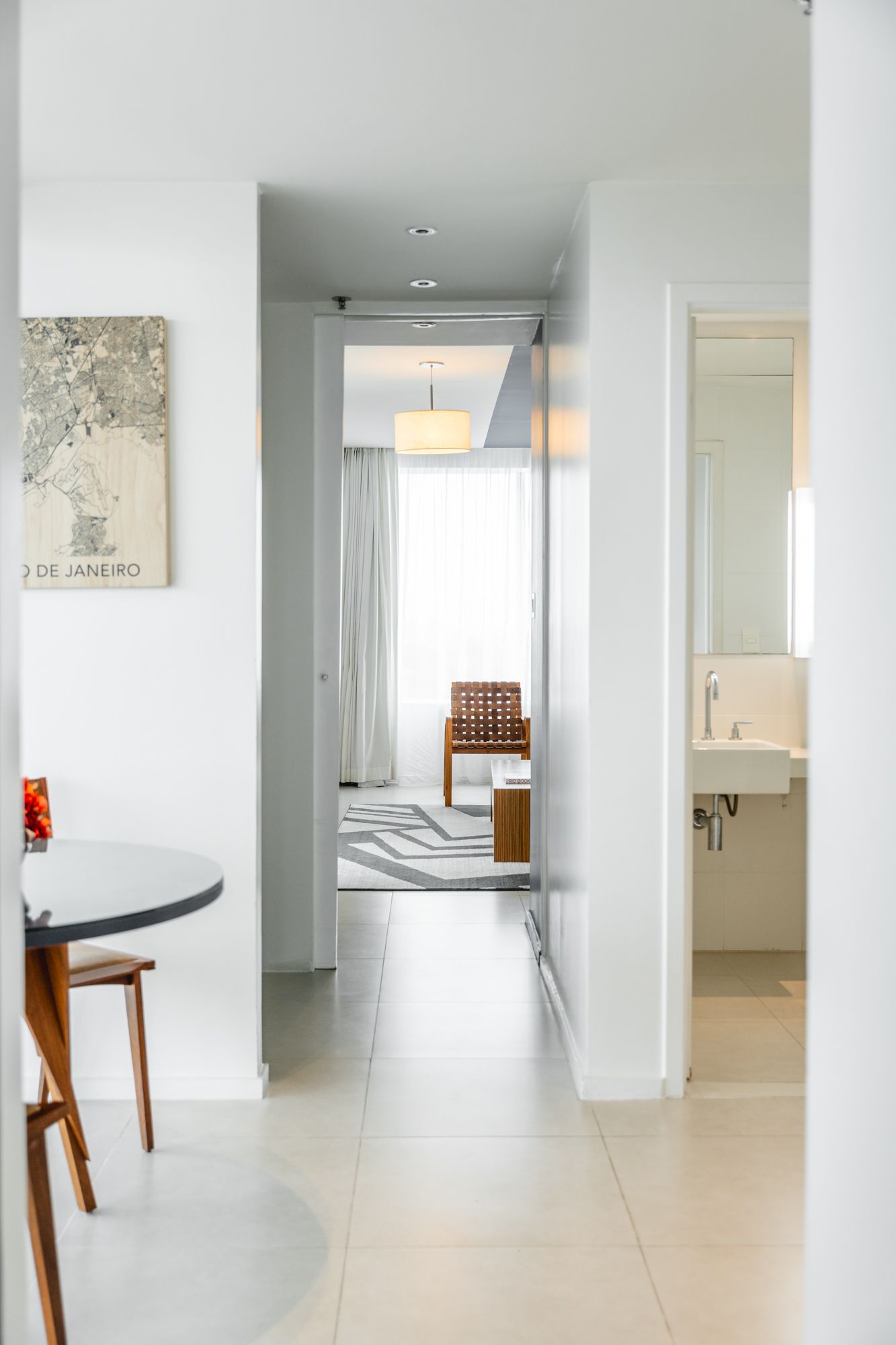 Corredor branco que leva a uma sala de estar, com um banheiro à direita. Uma mesa redonda com uma pintura pendurada à esquerda.